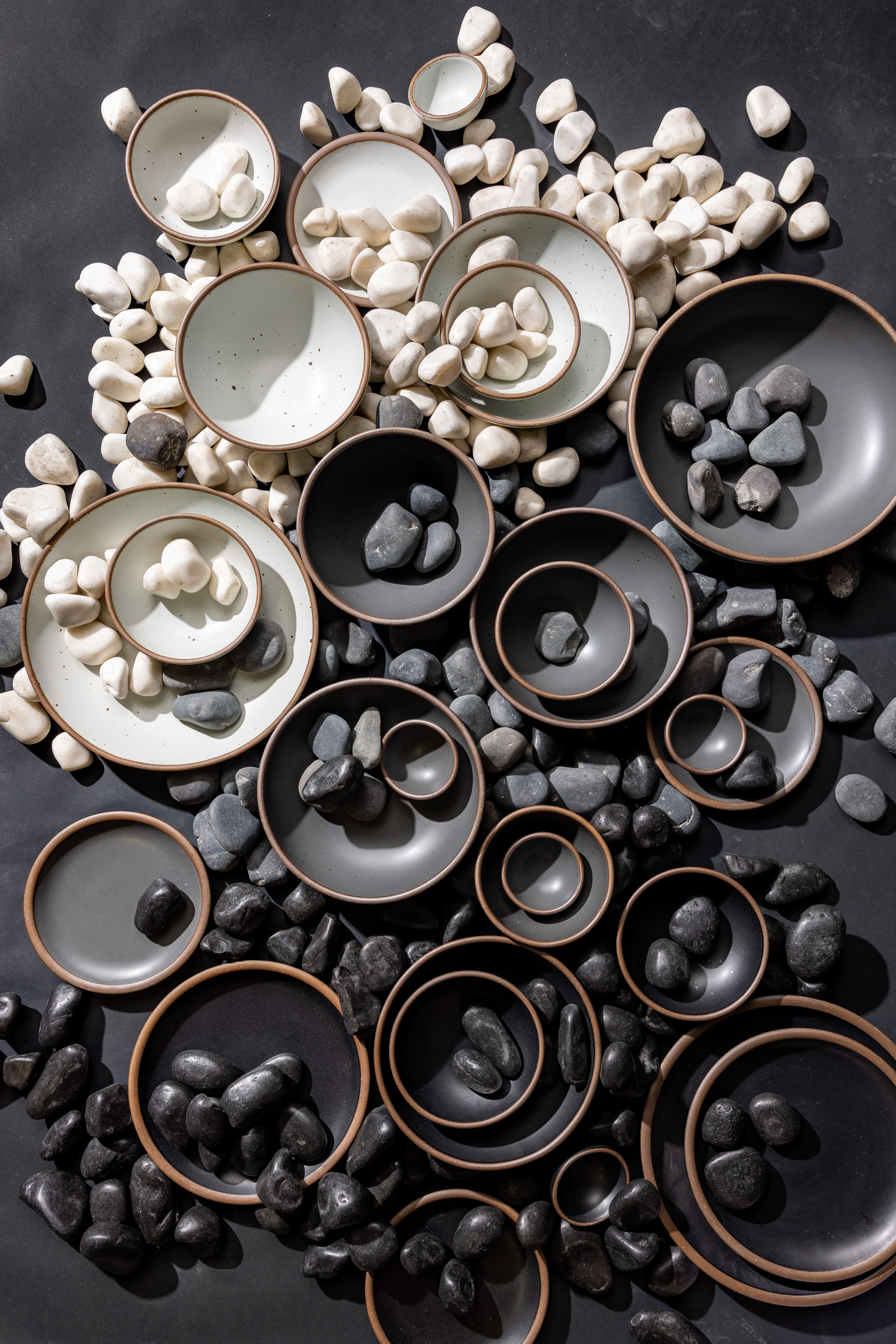 An overhead view of ceramic bowls and plates in various sizes in soft cool white, medium cool grey, and graphite - surrounded and filled by rocks of varying sizes and grayscale colors to match the pottery.