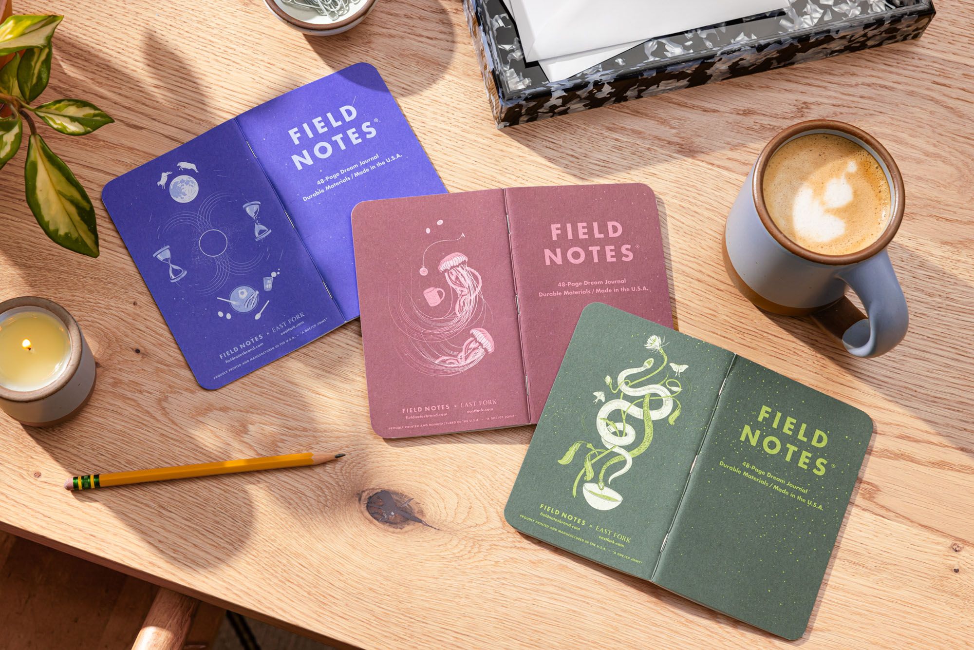 A 3 pack of Field Notes Journal are folded facing down on a desk featuring 3 illustrated back covers in a lapis blue, a plum, and an earthy green with a periwinkle mug filled with coffee