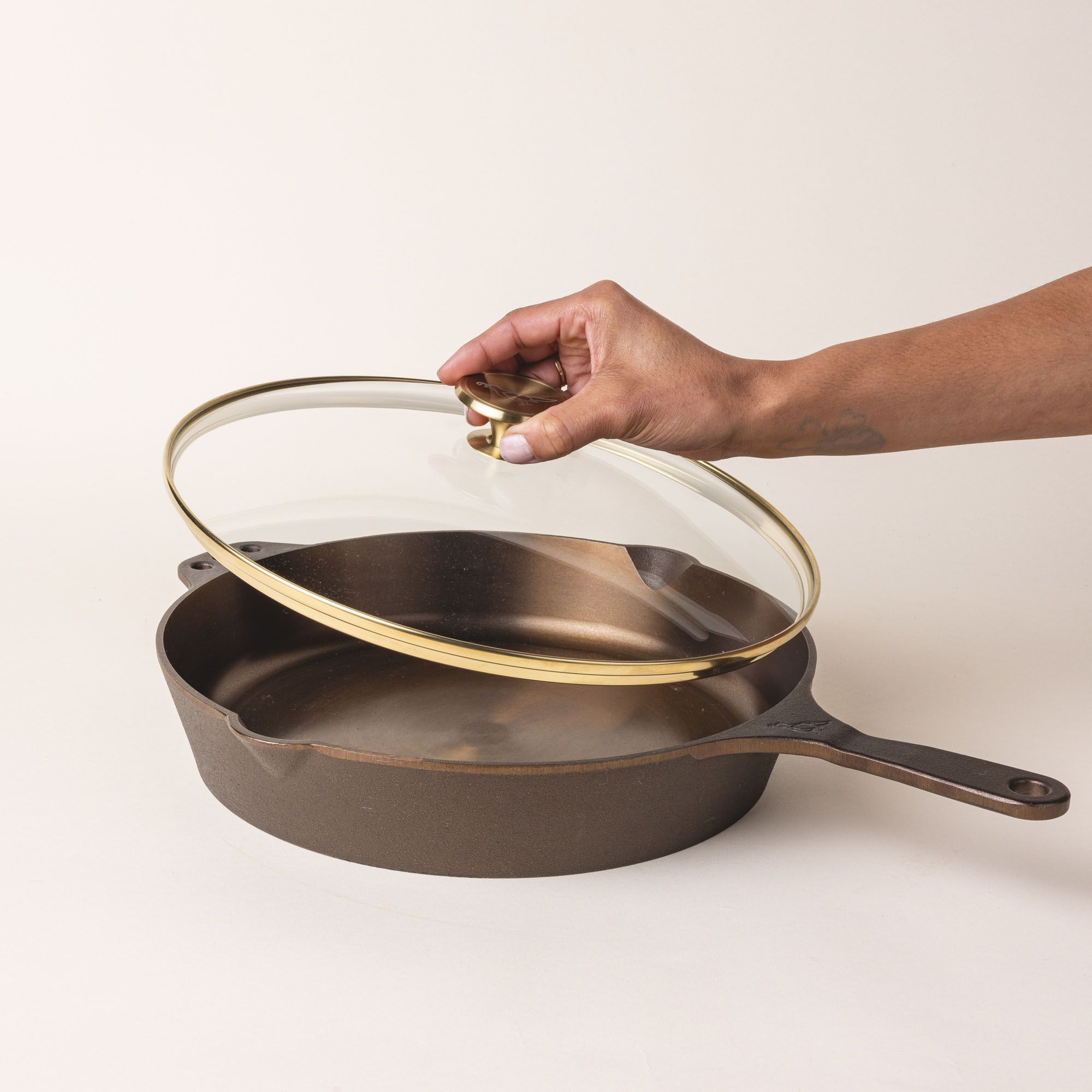 A hand holds up a glass skillet lid with a brass lip and brass handle knob over a cast iron skillet