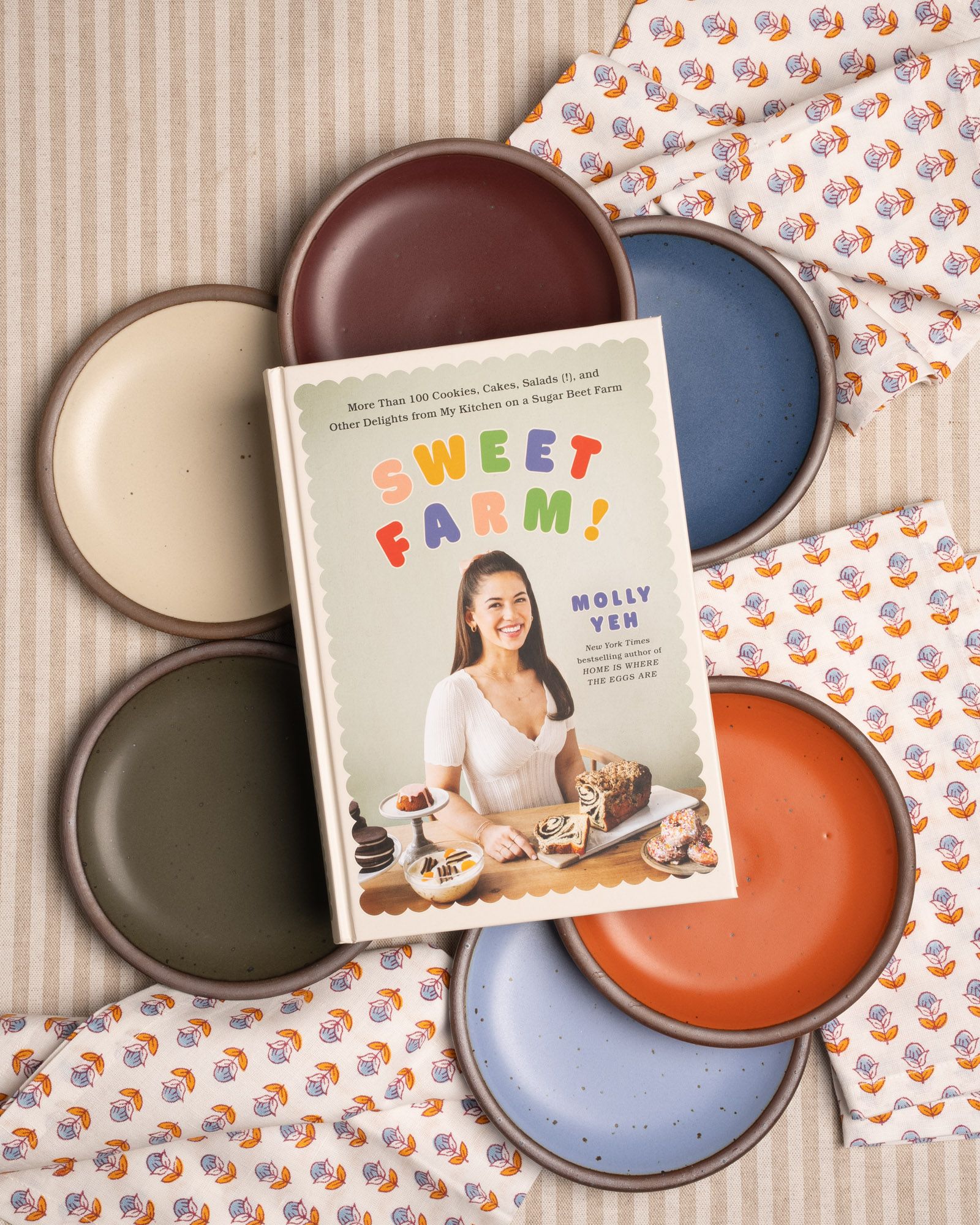 An overhead view of a cookbook titled 'Sweet Farm!' sitting on several colorful ceramic cake plates near periwinkle and amber flower napkins.