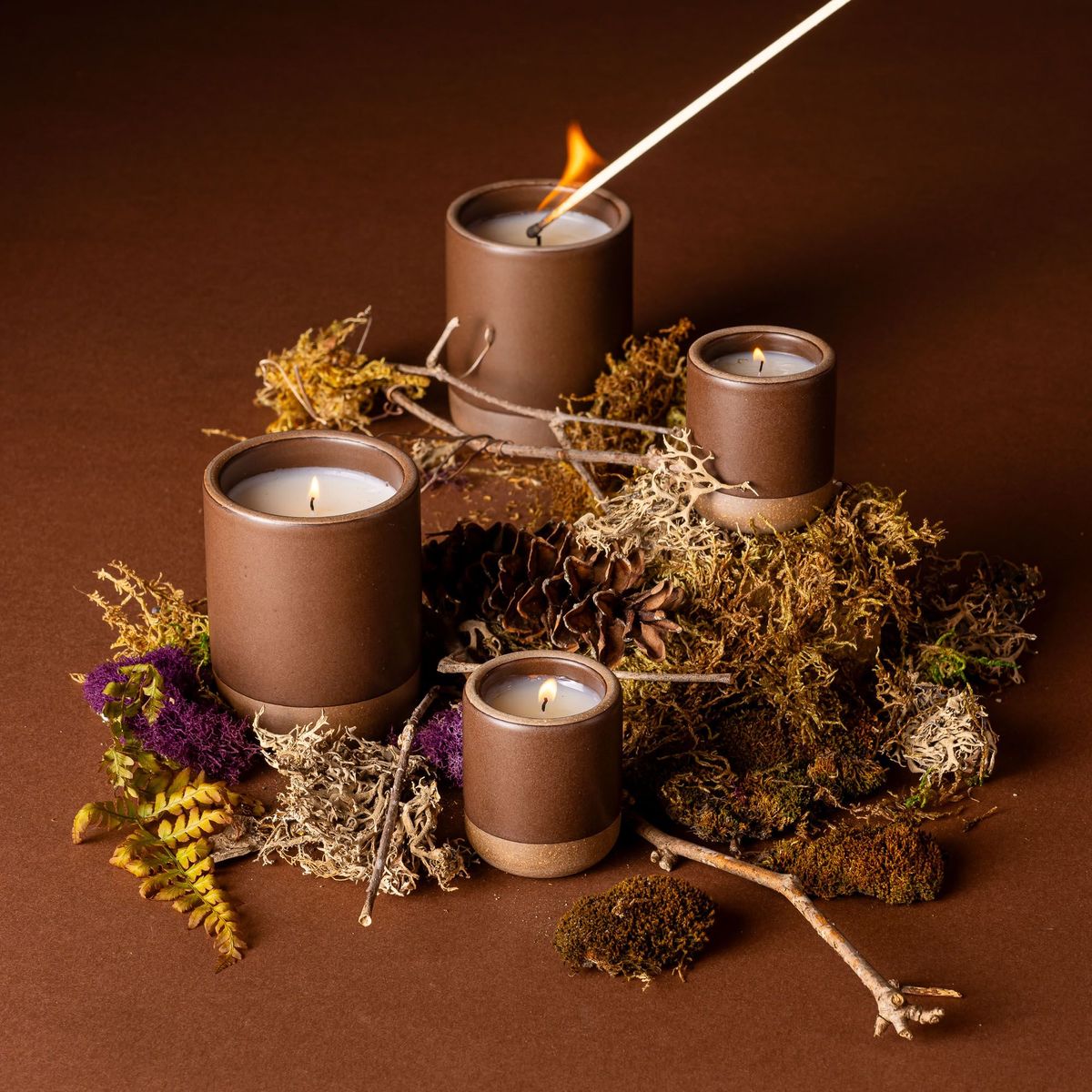 Four lit candles in ceramic vessels in a dark brown color with a match lighting one. They are artfully arranged and surrounded by dried grass, branches, and fern leaves.