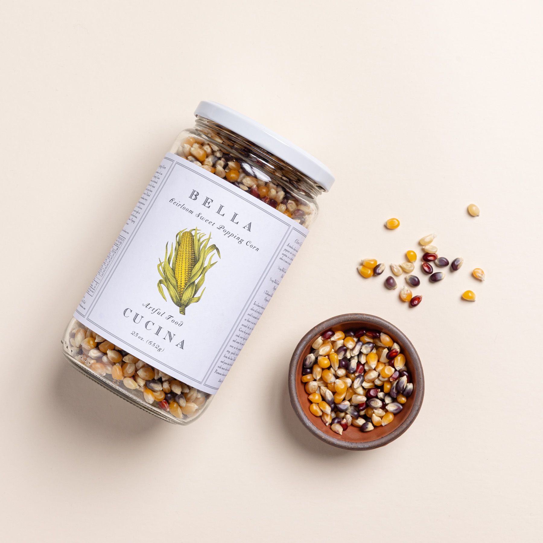 A jar with a white lid filled with popcorn kernels, and a large label with an illustration of corn and reads 'Bella Cucina Heirloom Sweet Popping Corn' next to a bitty bowl filled with kernels