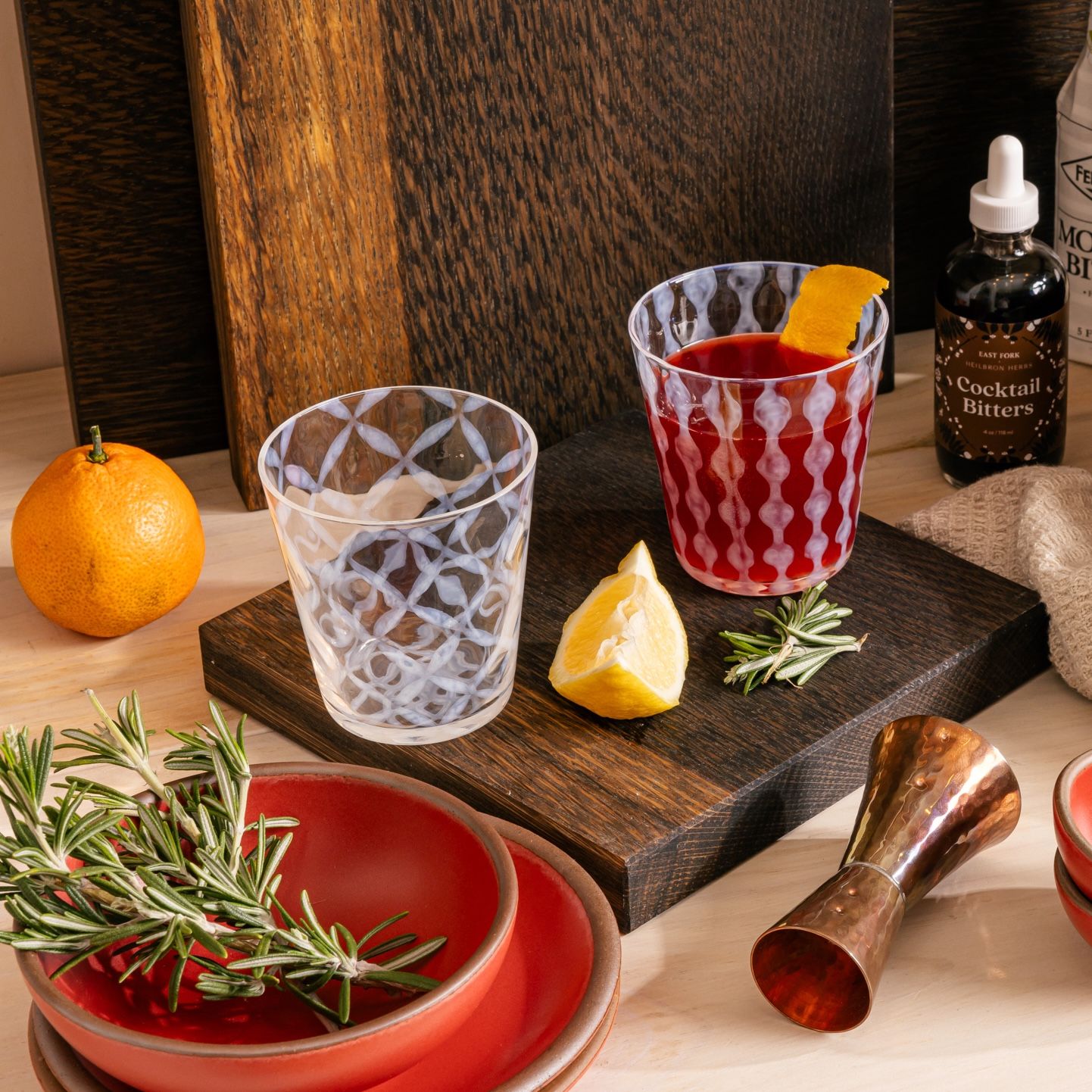 On a kitchen counter is a scene in the process of making cocktails, 2 tumbler glasses with frosted patterns, one filled with a red cocktail and orange slice. There's also walnut cutting boards, rosemary twigs, lemon slice, a shot measurer, and a small bowl and plate in a bold red color.