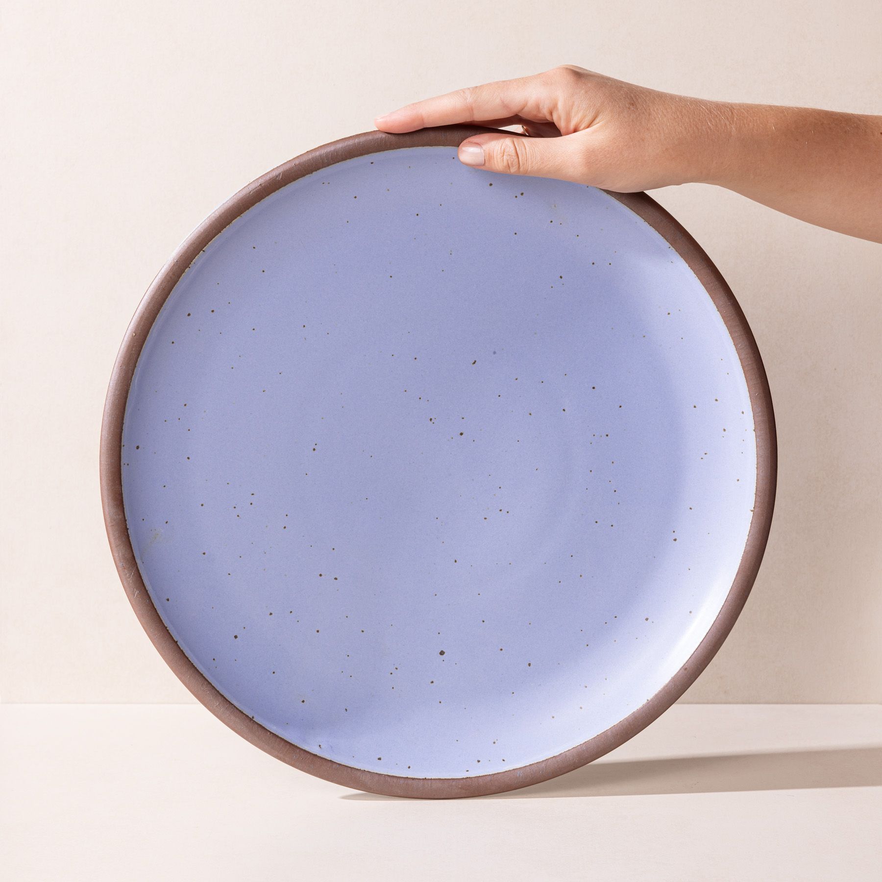 A hand props up a large ceramic platter in a periwinkle color featuring iron speckles and an unglazed rim