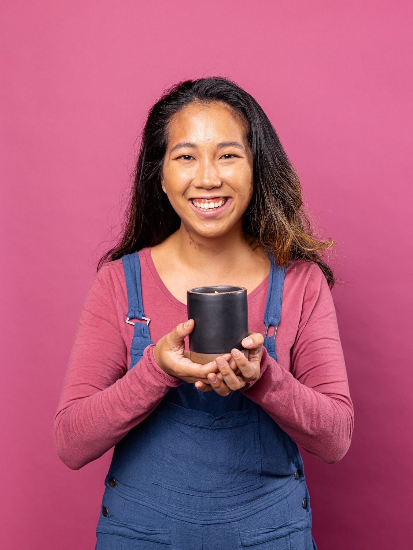 A person smiles in overalls and holding a large lit candle in a ceramic vessel against a pink background