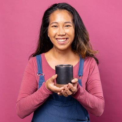 A person smiles in overalls and holding a large lit candle in a ceramic vessel against a pink background