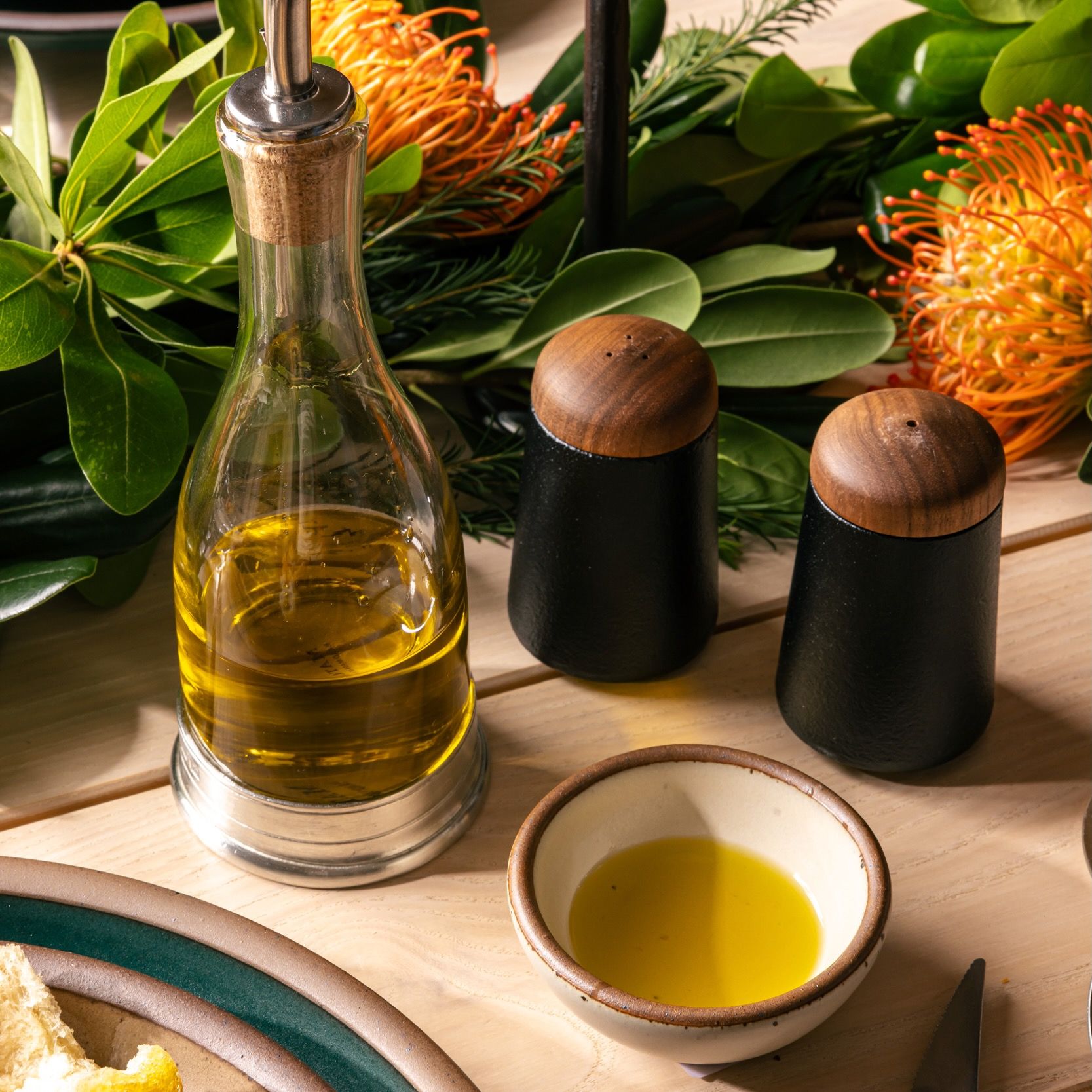 On a table, sits a set of salt and pepper shakers with a black cast iron base with a rounded walnut wood top on a white background. Also on the table is greenery centerpiece, a glass and pewter oil cruet, and a tiny white bowl filled with oil.