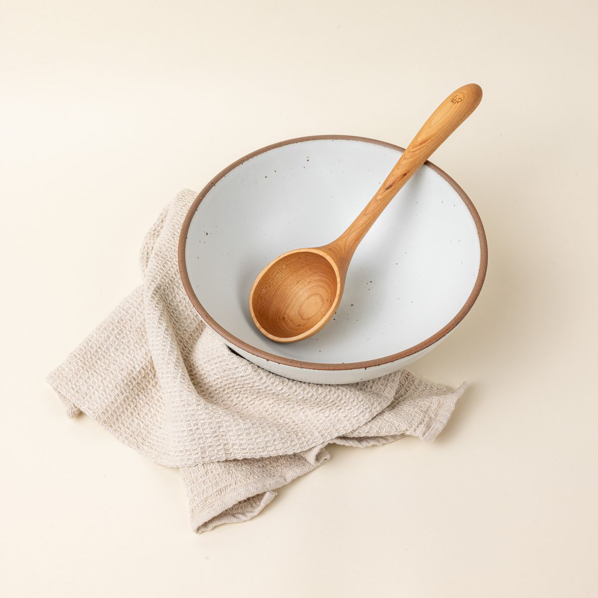 A wooden ladle with a large scoop sits inside a large white ceramic mixing bowl with a cream towel.
