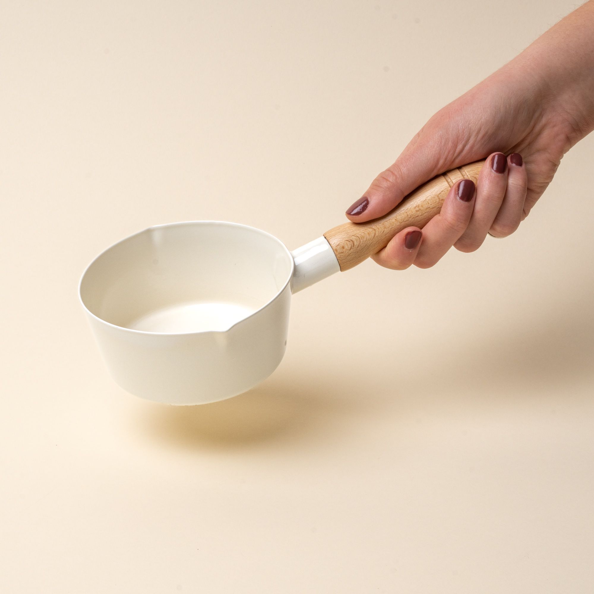 A hand holds a white enamel pot with simple spouts on the side with a wooden handle