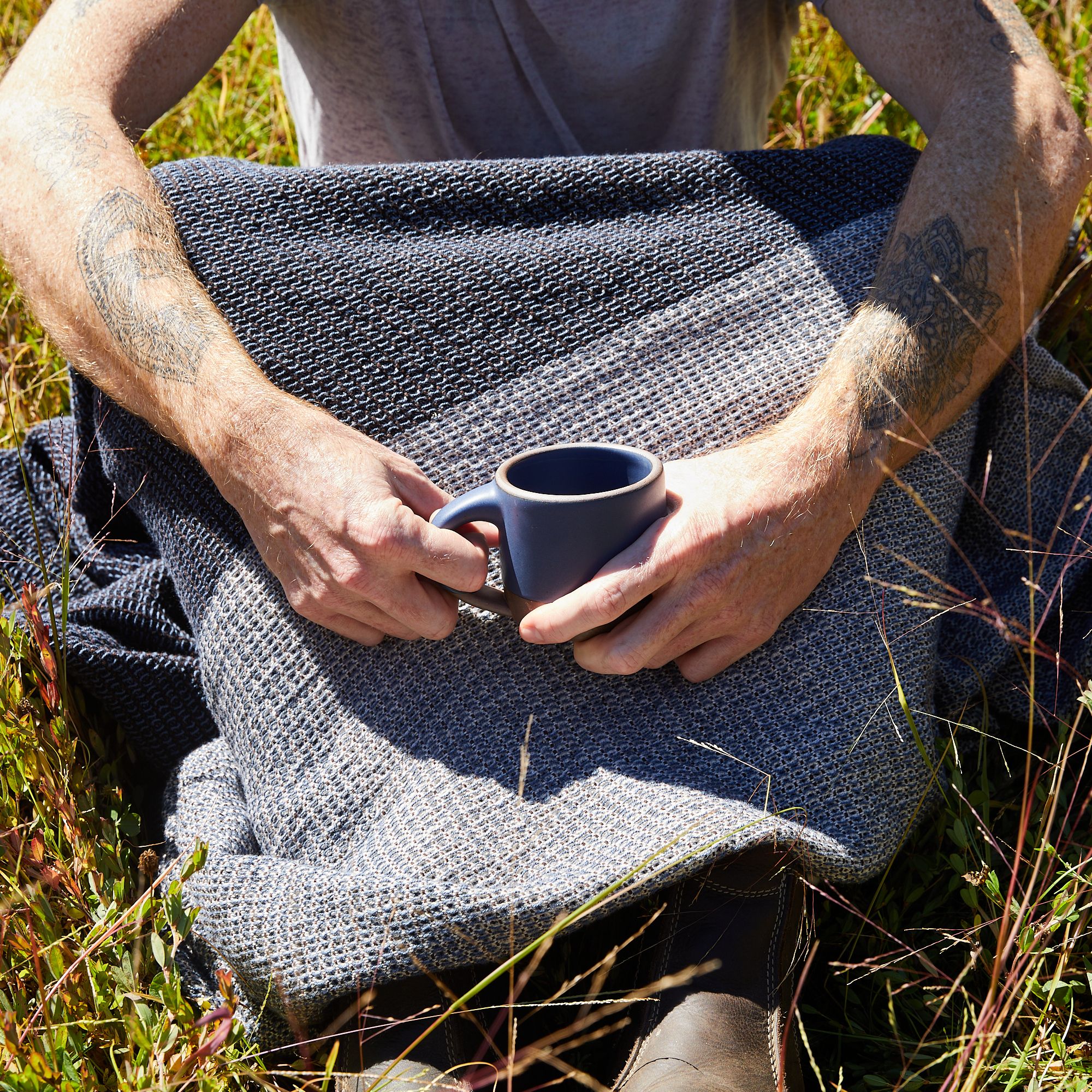 A person with a blanket over their lap, shown chest to feet, sitting in tall grass holding a blue small mug with both hands in front of them