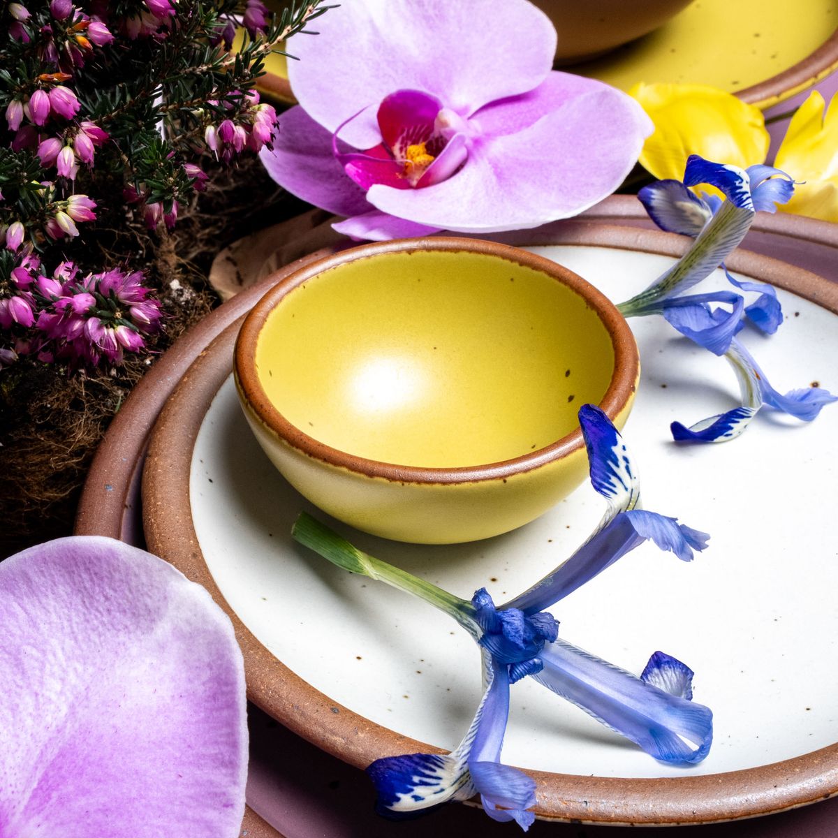 A tiny rounded ceramic bowl in a zesty yellow color featuring iron speckles and an unglazed rim, sitting on a white plate surrounded by flowers.