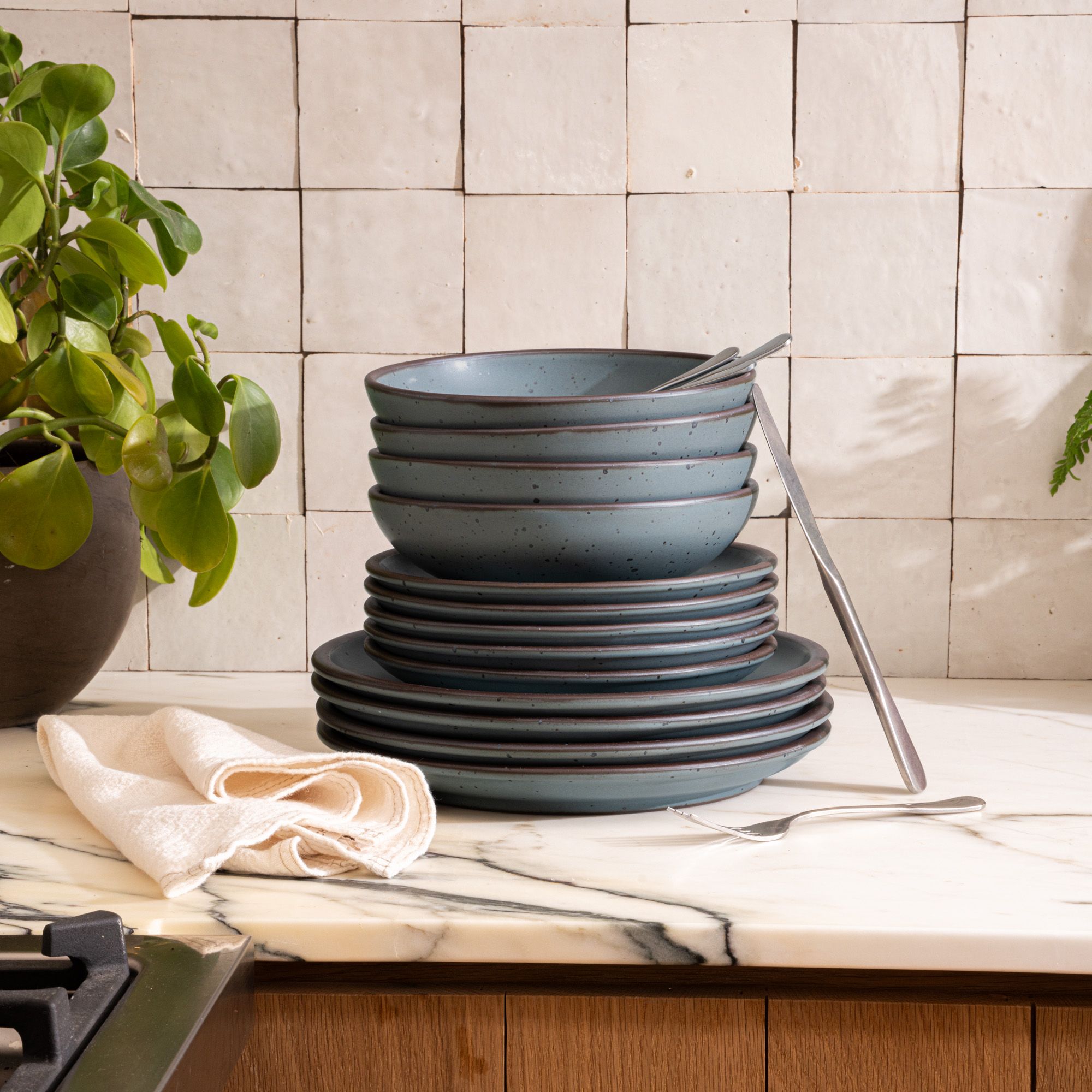 4 ceramic dinner plates, 4 side plates, and 4 shallow dinner bowls in a blue-grey color with iron speckles and unglazed rims are stacked all together on a countertop with a napkin and plant