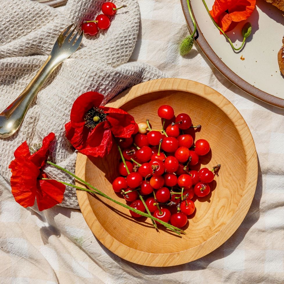 A light wood bowl is filled with fresh cherries and poppy stems, to the left is a brass picnic spork. All are sitting on a cream tablecloth and kitchen towel.
