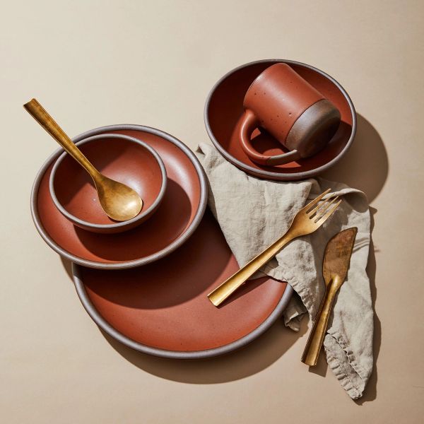 An artful arrangement of ceramic plates, bowls, and a mug in a cool terracotta color, with a linen napkin and brass fork, spoon, and knife.