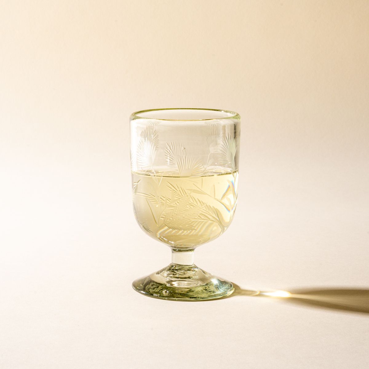 A clear short wine glass made of recycled glass with etched thistle designs on it, filled with white wine.