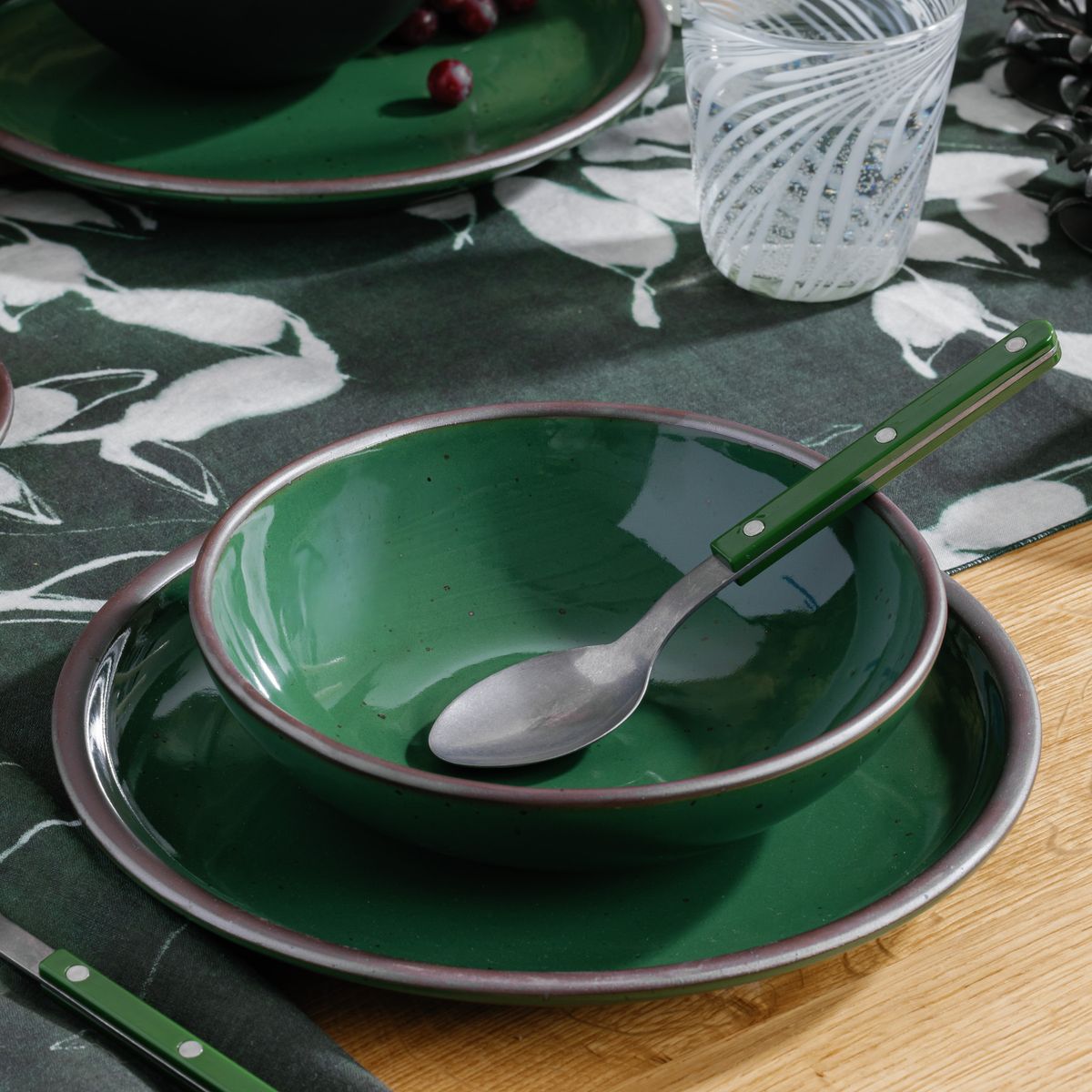 A glossy emerald green ceramic bowl stacked on a matching plate with a spoon resting inside, set on a wooden table with a dark green patterned runner.