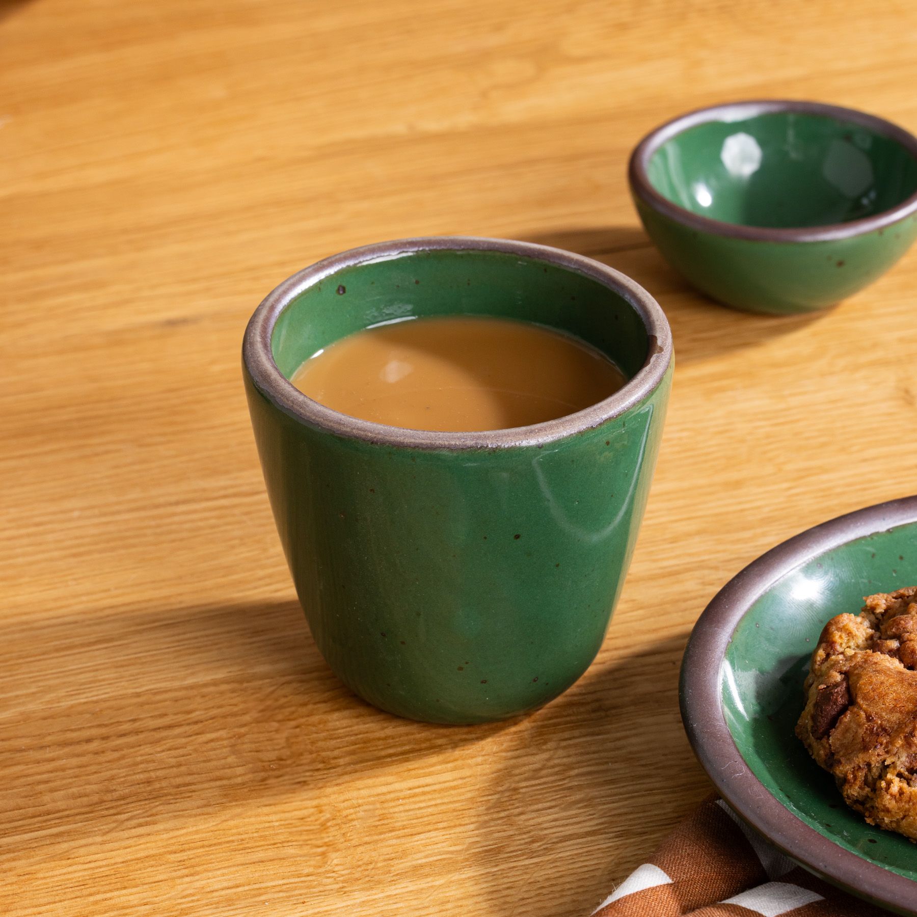 Emerald green ceramic cup filled with coffee with cream, placed on a wooden table beside a cookie on a matching plate.