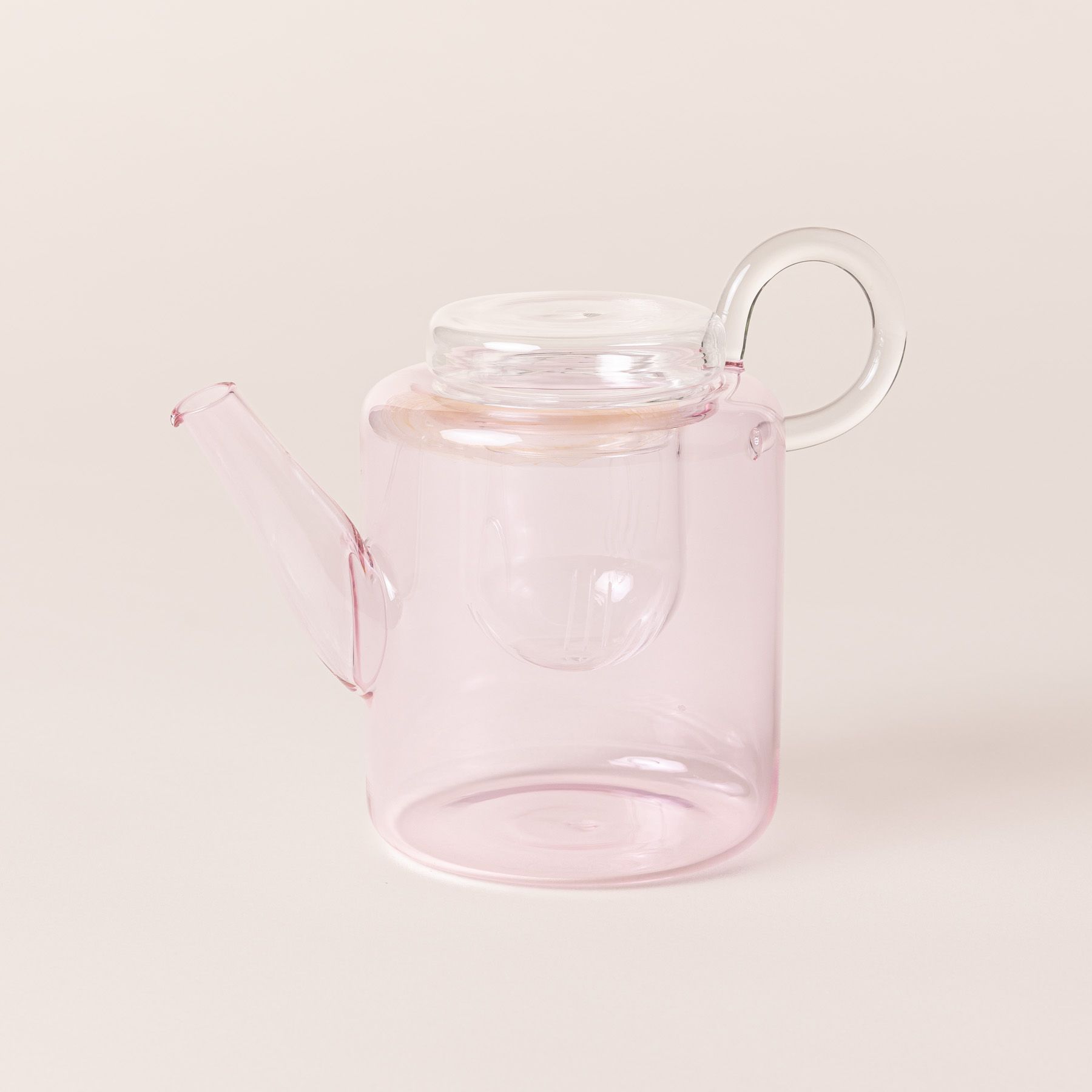 A hand holds a modern glass teapot in a faint pink color with a strainer inside