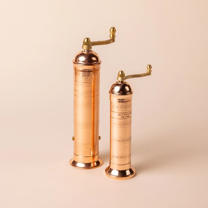 Two copper pepper and salt mills together. Both have a flanged base and a rotating grinder on top. The pepper mill is taller and slightly wider.