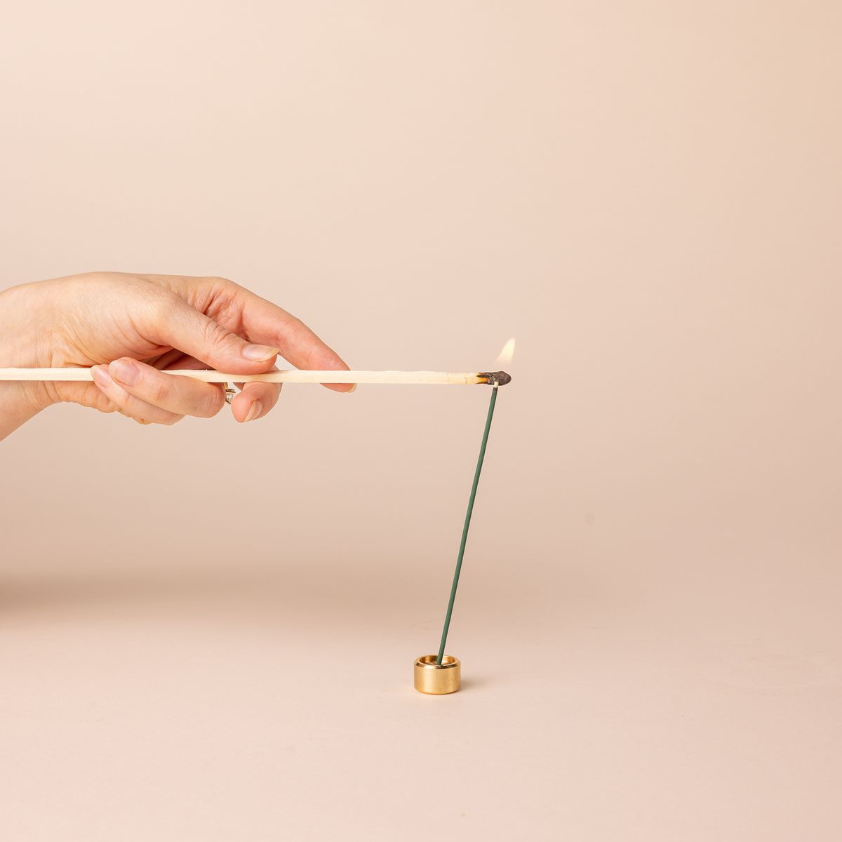 A hand holding a match lighting an incense stick. The incense stick sits in a short small brass cylinder incense holder.