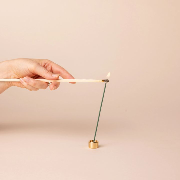 A hand holding a match lighting an incense stick. The incense stick sits in a short small brass cylinder incense holder.