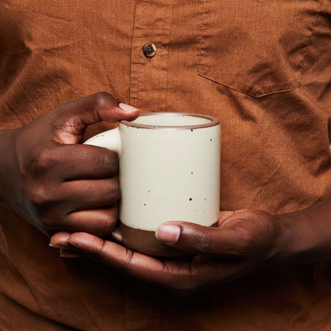 A hand holds an East Fork mug in Panna Cotta, an off-white color featured iron speckles.