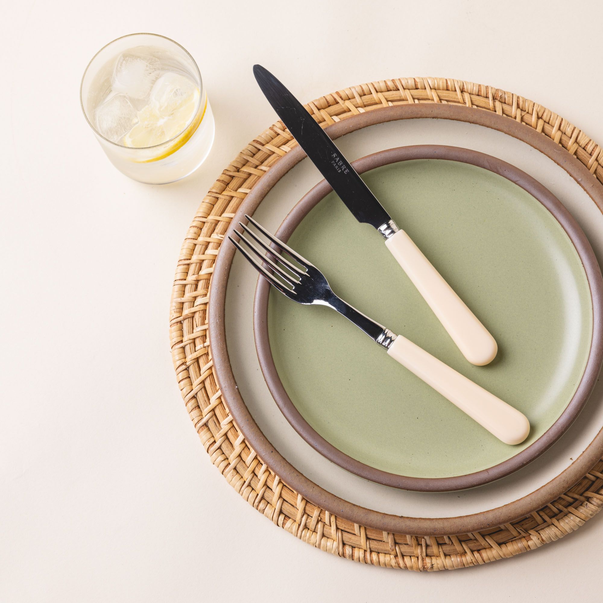 A place setting with 2 ceramics plates, 1 sage green, and 1 neutral, sitting on a woven natural charger. A cream matte handled fork and knife are arranged on top of the plates.
