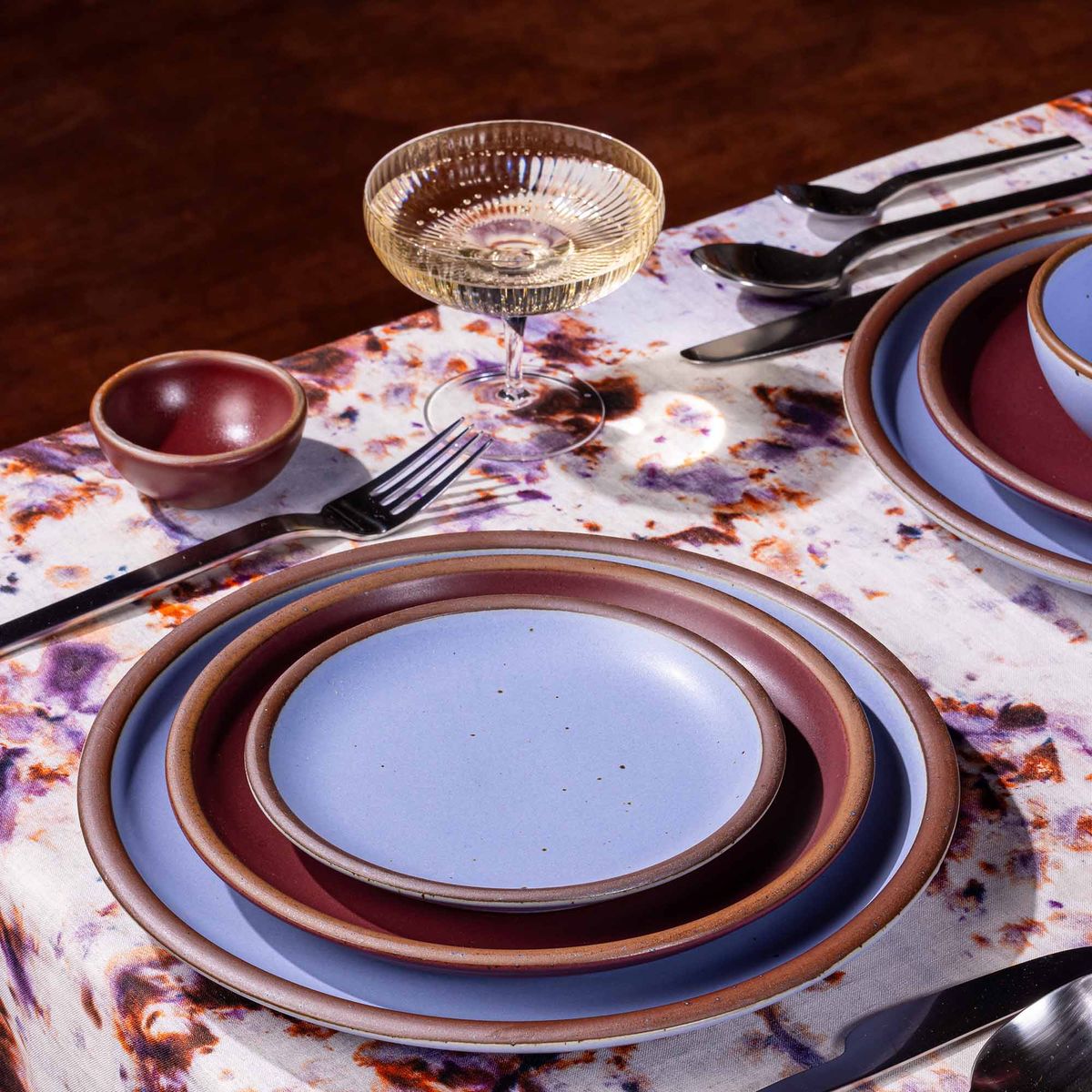 A table setting featuring marbled textiles, a coupe glass, and plates in plum and periwinkle colors