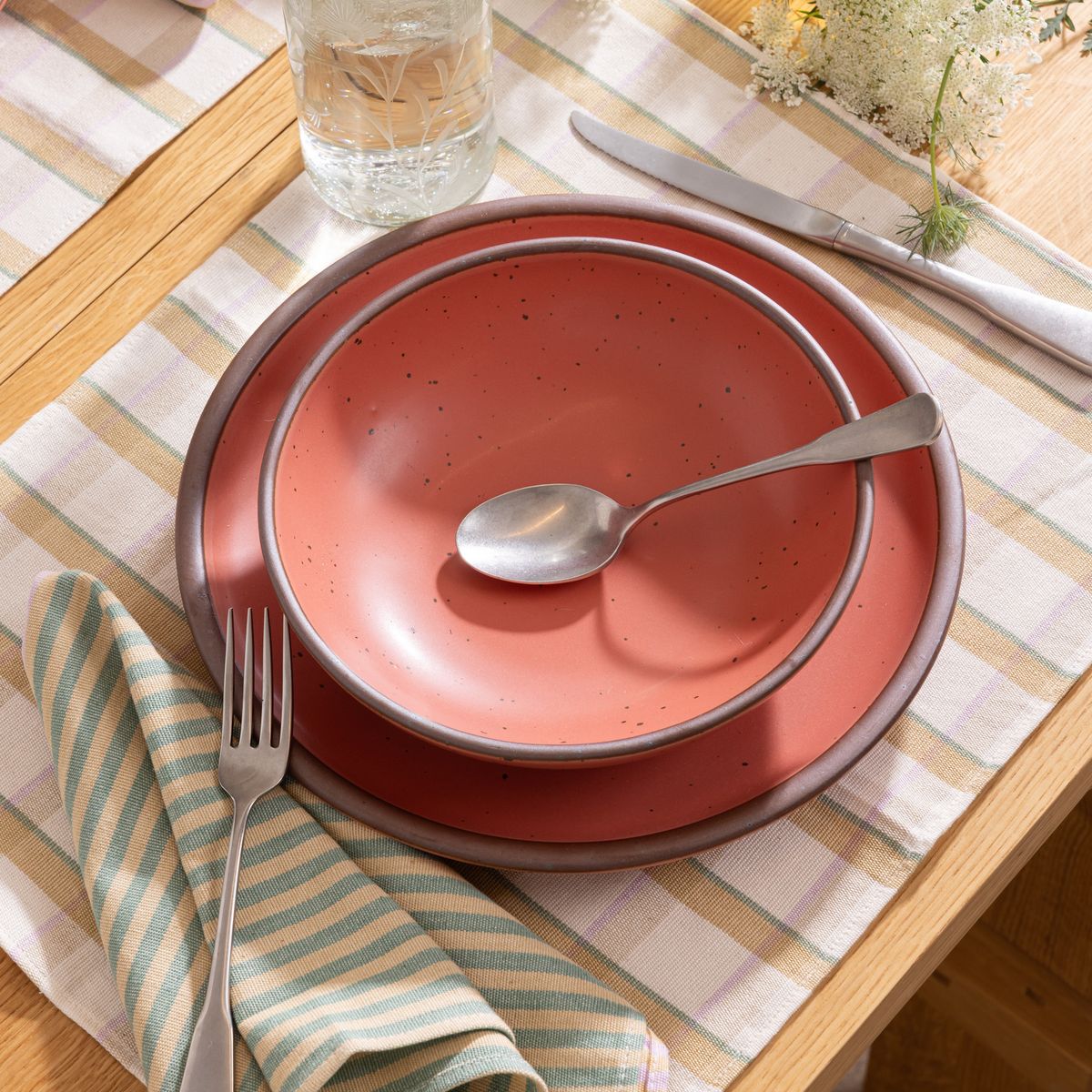 Watermelon pink stoneware shallow bowl and dinner plate stacked with silverware on a plaid placemat, striped napkin, and glass of water nearby.