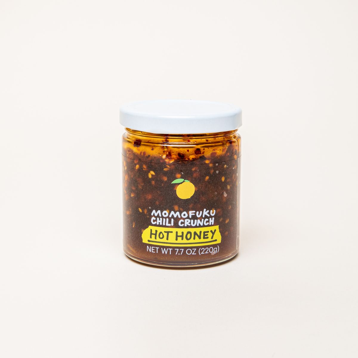A clear jar with a white top that is filled with a warm brown honey with chili flakes and a label that reads, "Momofuku Chili Crunch Hot Honey"
