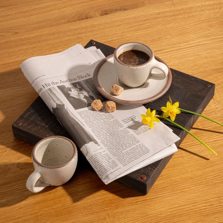 Small stoneware espresso cup with handle and saucer, in a warm off-white color with iron speckles, styled with another espresso cup, newspaper, and flower stems.