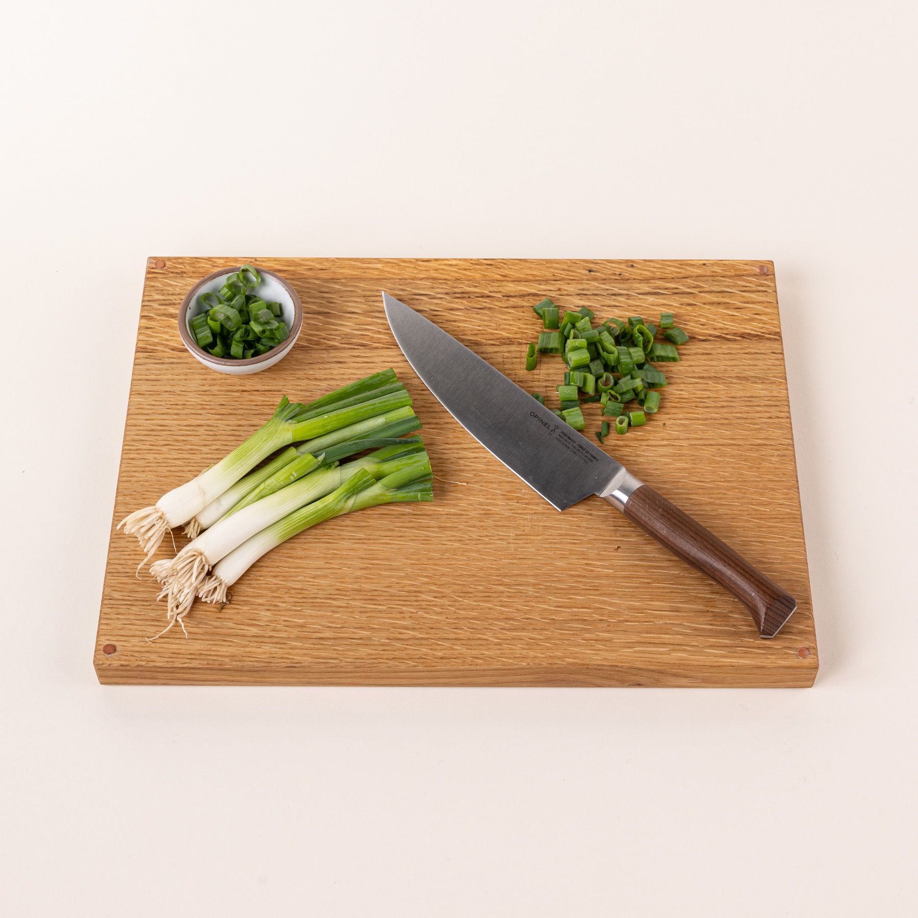 On a cutting board, there's a chef's knife with a sharp steel blade with a beveled dark wood handle and chopped green onions