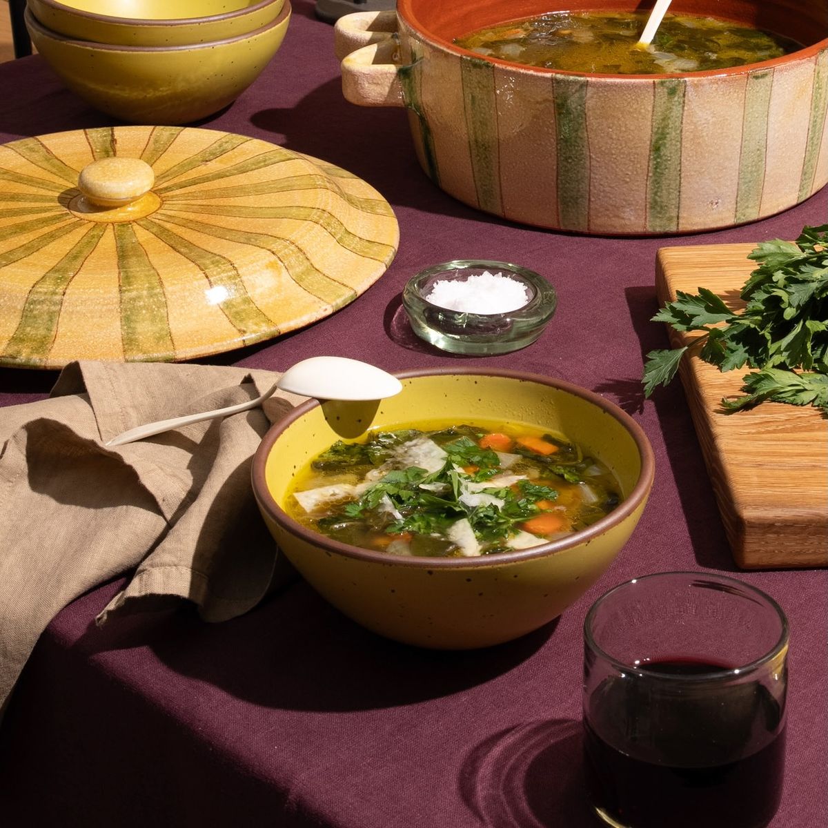 A ceramic soup bowl in a zesty yellow color, filled with soup, and with an enamel spoon, casserole dish, ingredients, a drink, and a purple tablecloth