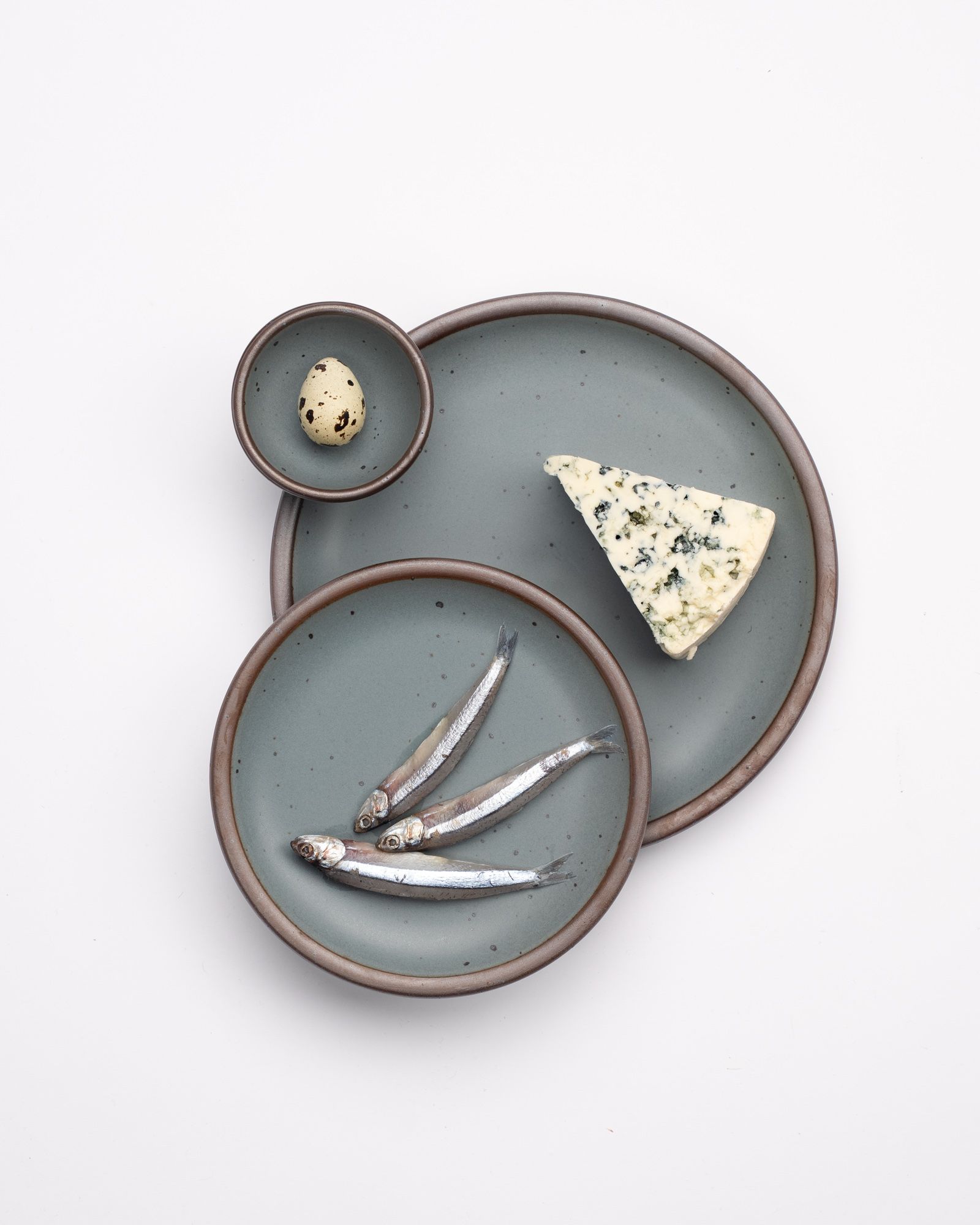 An overhead view a ceramic dinner plate, side plate and tiny bowl in blue-grey colors with styled food on each including fish, cheese, and a quail egg.