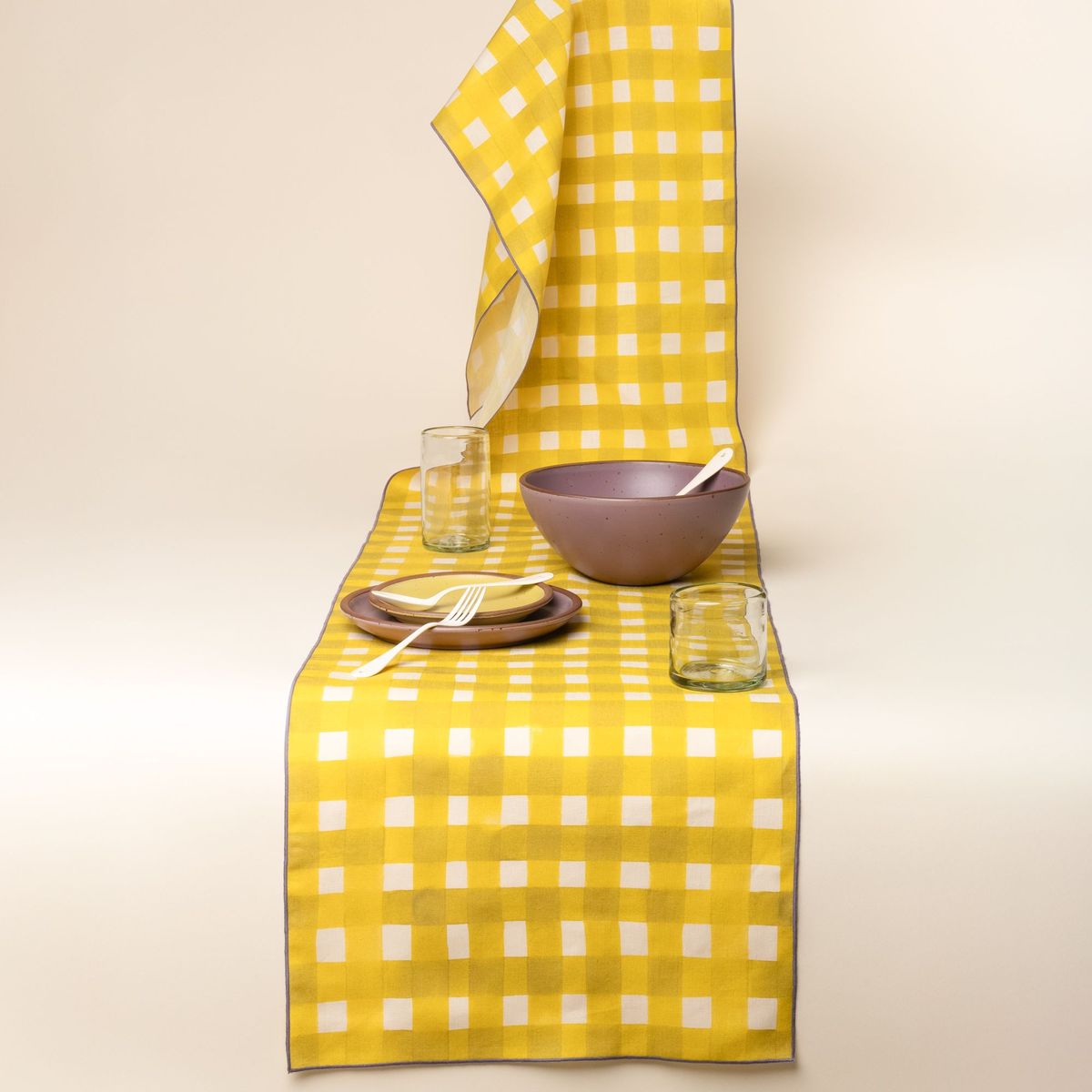 A purple plate and bowl with glasses sit on a table runner featuring a bright yellow crosshatch pattern with purple edges.