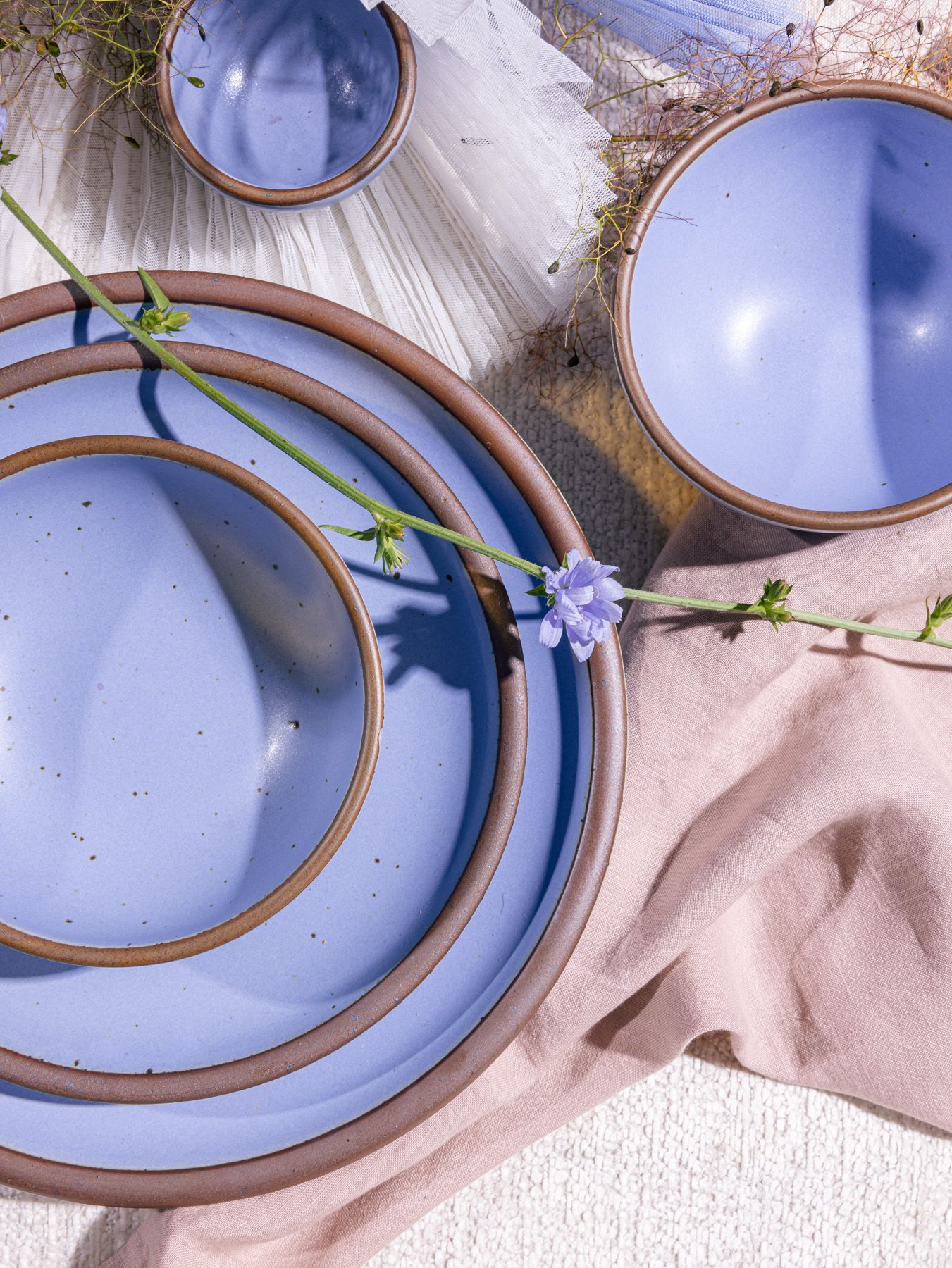 Artistically styled stack of periwinkle ceramic dinnerware plates and bowls on pale pink table cloth, with a single flower spring laying across them. 