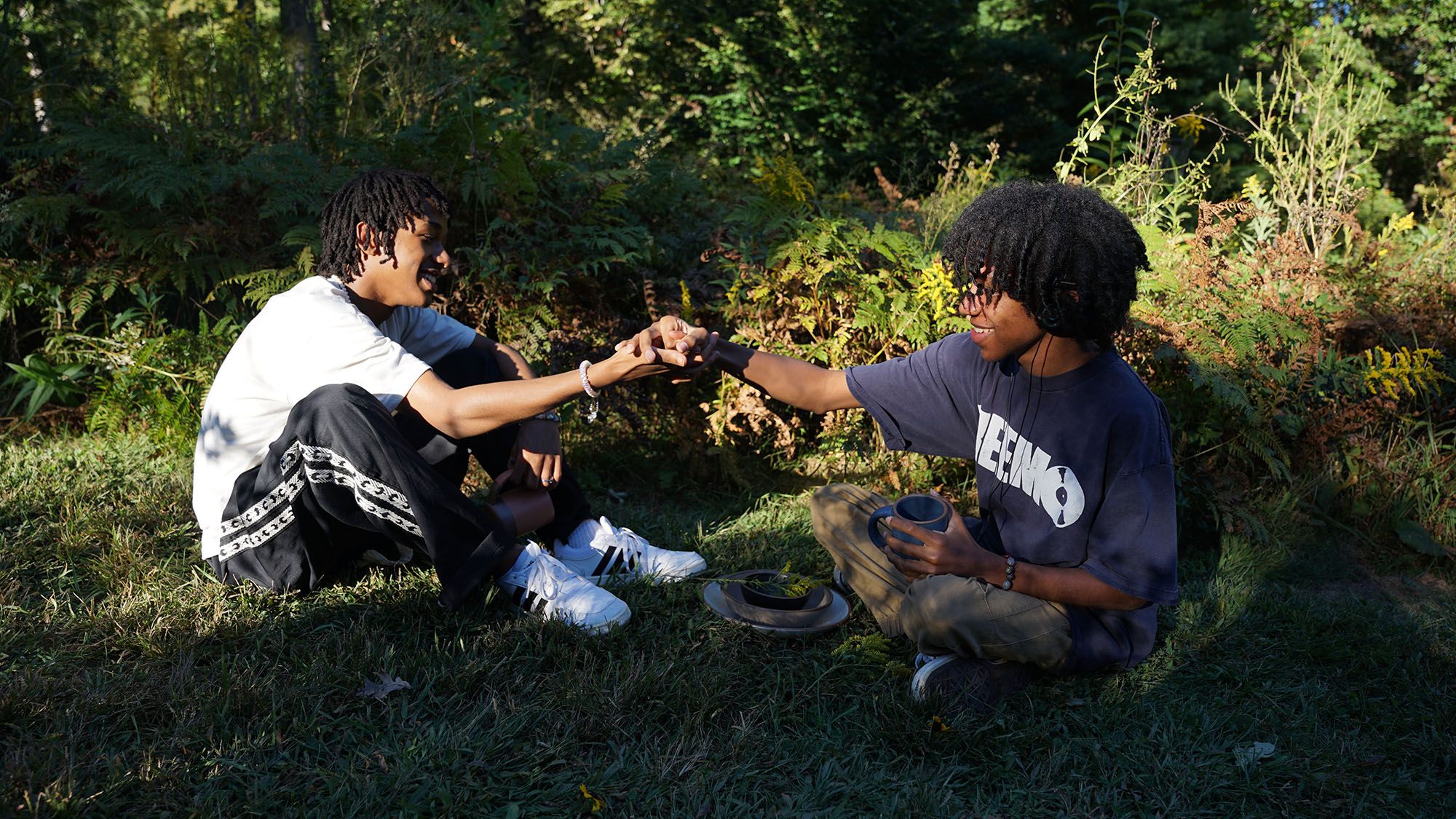 Two kids share a handshake while sitting down outside surrounded by woods