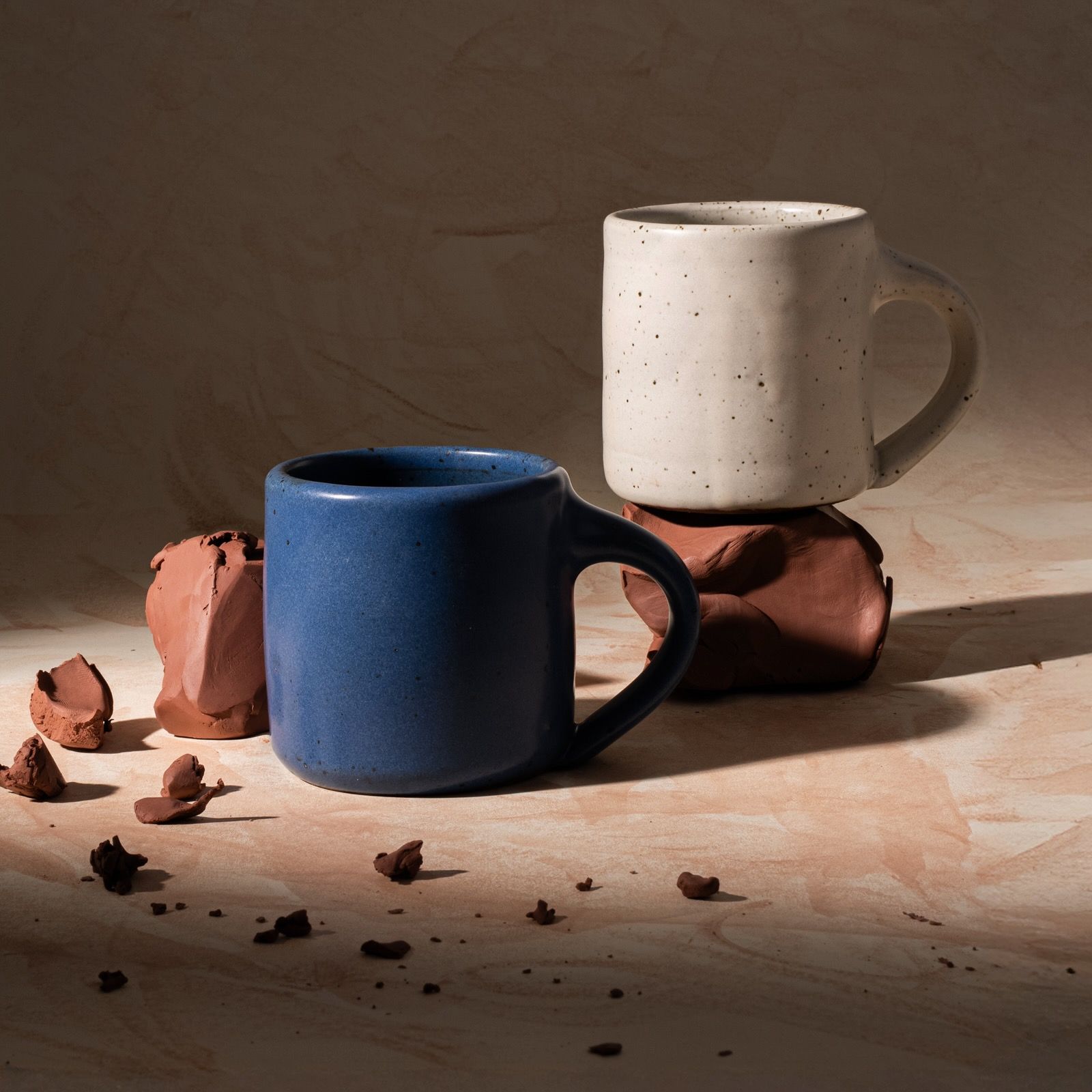 Two medium sized ceramic mugs with handles in a warm, tan-toned, off-white and cool, medium blue colors featuring iron speckles, styled with raw clay