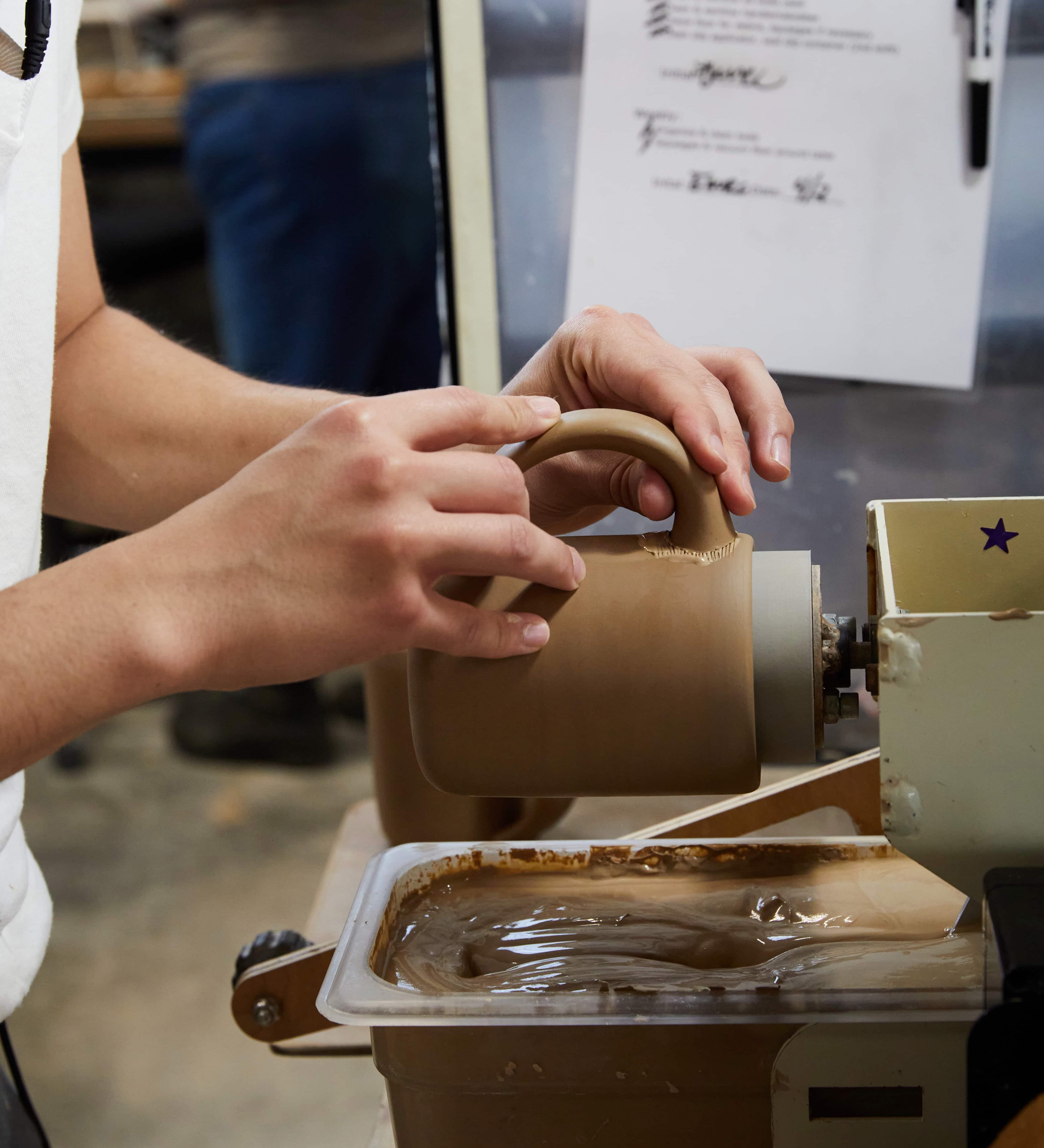 A hand is attaching a handle on a ceramic mug in a factory setting.