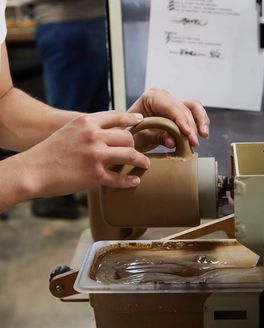 A hand is attaching a handle on a ceramic mug in a factory setting.
