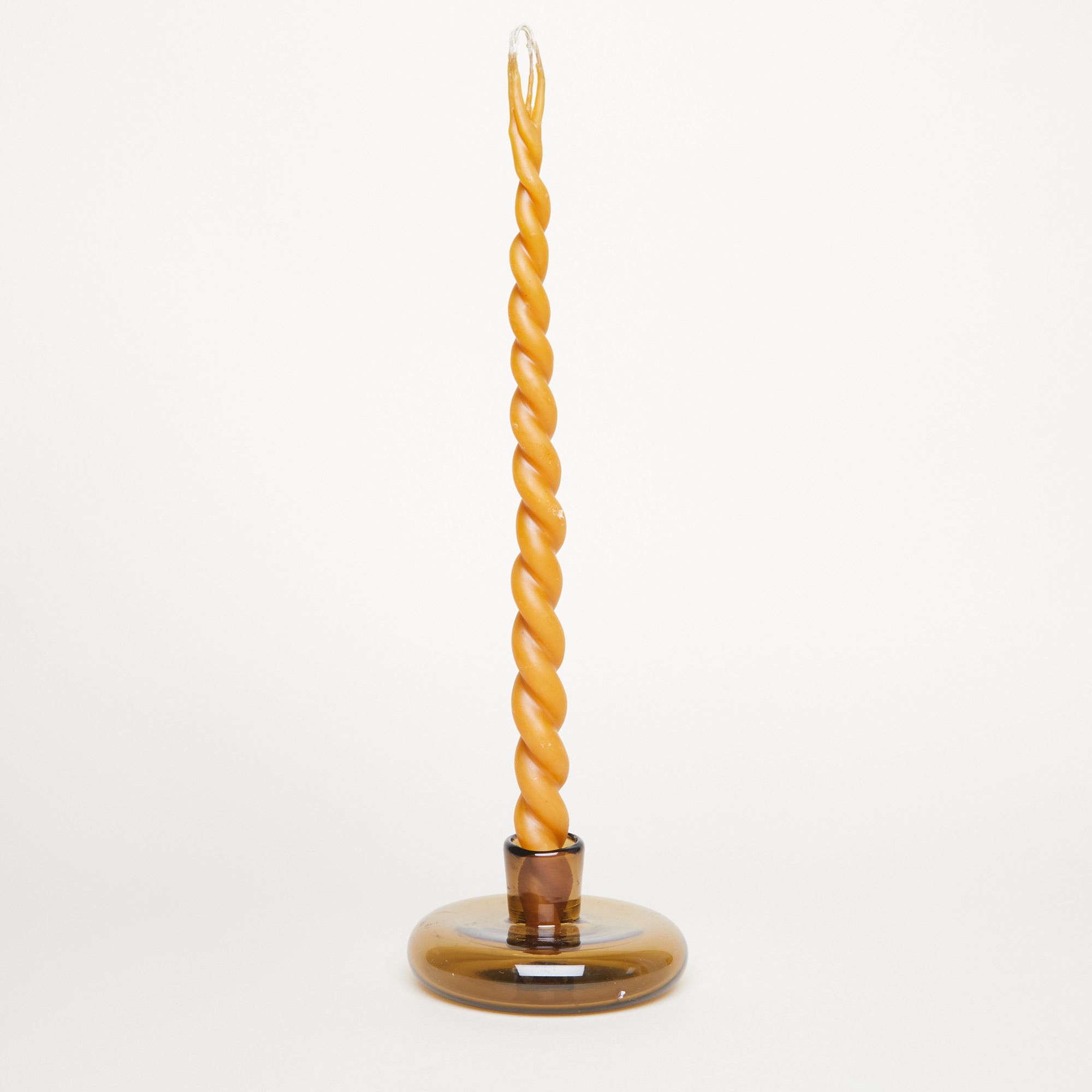A long, yellow twisted wax candle with a double wick stands in a clear brown circular candle holder