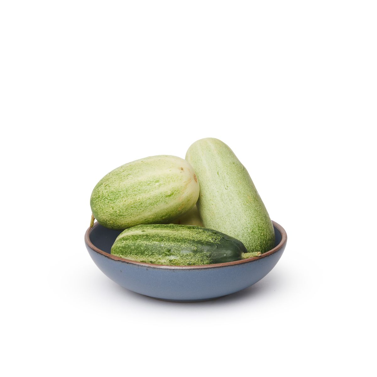 A dinner-sized shallow ceramic bowl in a cool, medium blue color featuring iron speckles and an unglazed rim, filled with cucumbers