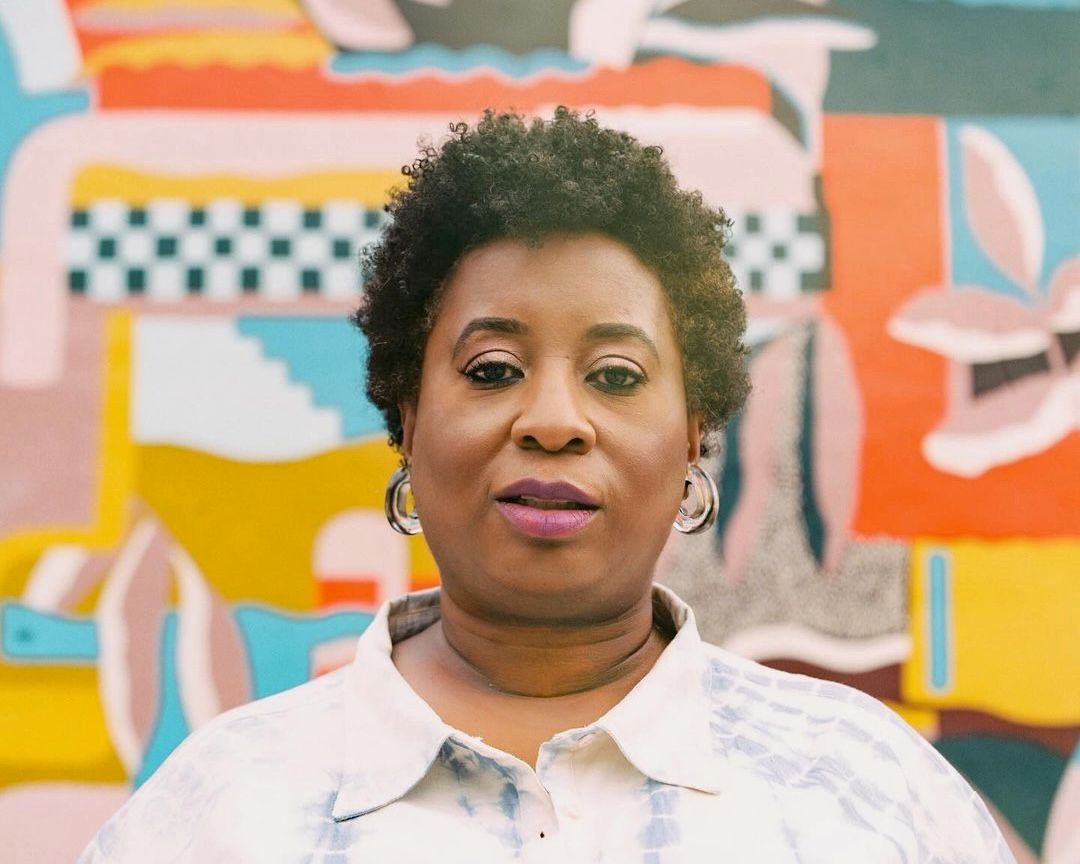 A woman in a collared shirt and large glass earrings stairs at the camera, behind her is a colorful mural.