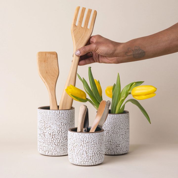 Three white ceramic vessels in Small, Medium, and Big with cracks and the interior being dark teal. Each vessel is filled with wooden utensils, flowers, flatware.