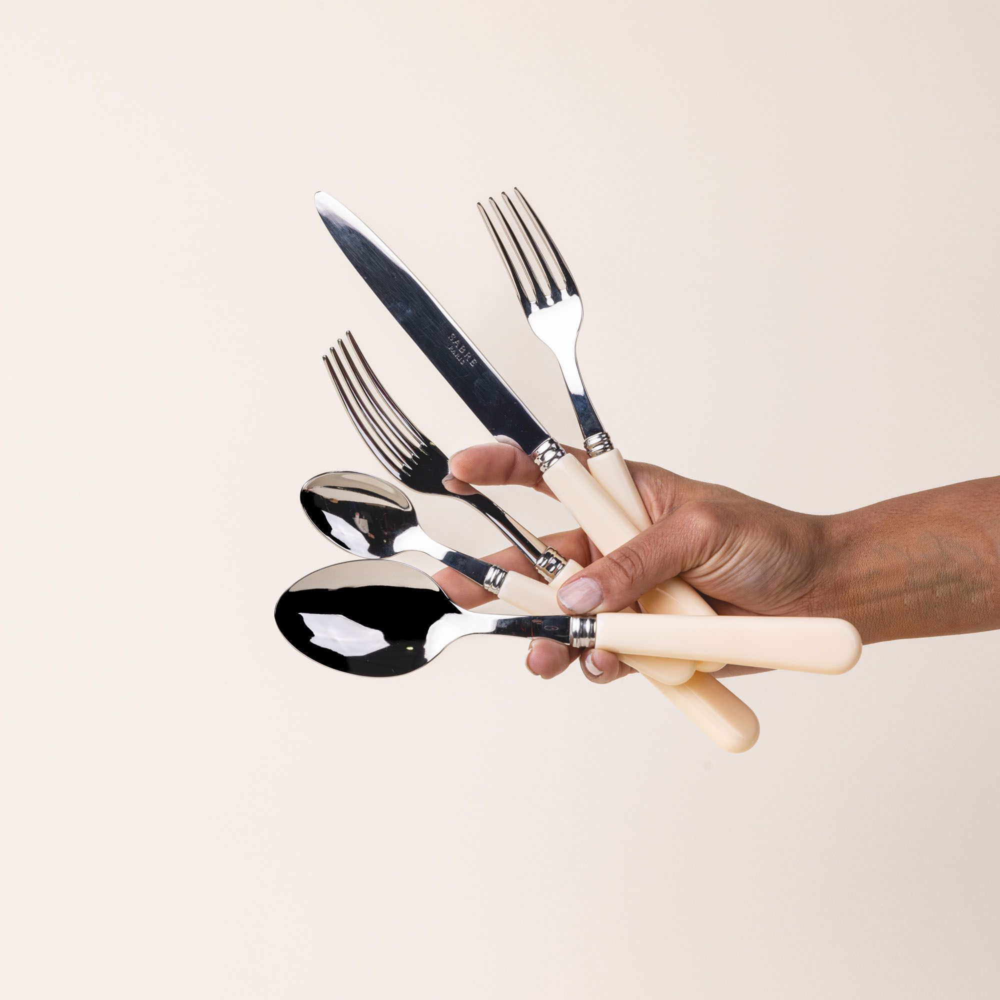 A hand holds a five piece flatware set with shiny utensils and matte cream handles