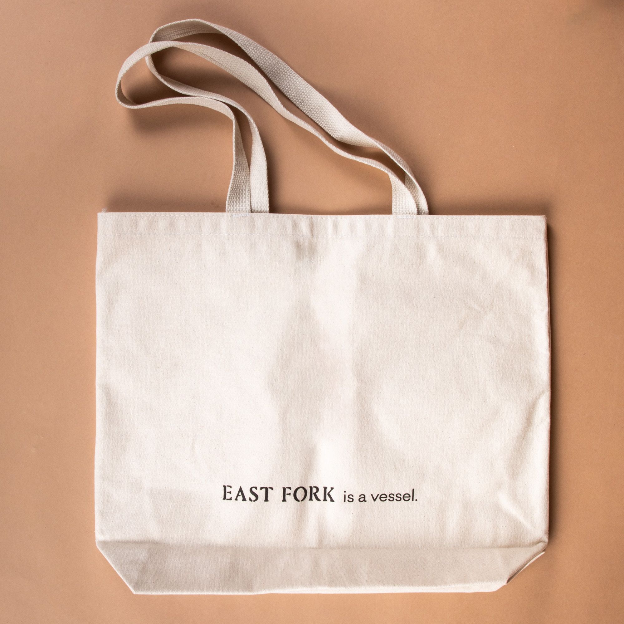 Person holding a cream canvas tote on their shoulder with their hand holding onto the handle - the tote has a charcoal illustration on the front of a bowl with fruit, little bowls, plate, and a mug. Underneath, it reads 'East Fork is a vessel'.