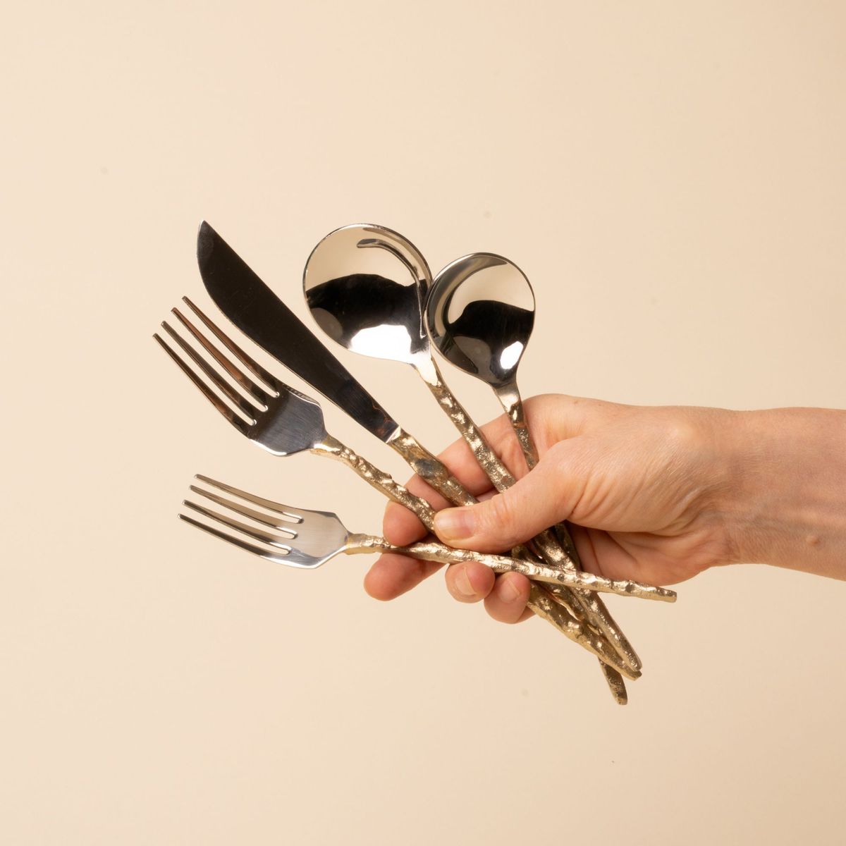 A hand holds a shiny silver flatware set with hammered gold handles arranged on a beige background.