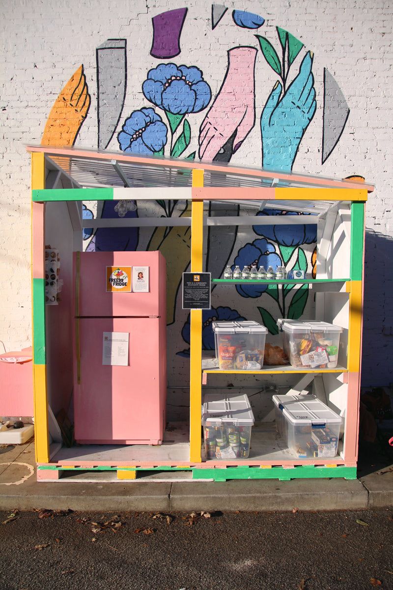 In front of a colorful outdoor mural is a structure with bins of food, plastic water bottles, and pink refrigerator.