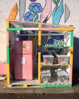 In front of a colorful outdoor mural is a structure with bins of food, plastic water bottles, and pink refrigerator.