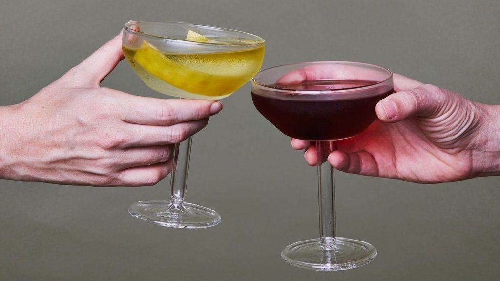 Two hands each hold a Martini coupe, one filled with clear liquid and a twist of lemon and the other filled with red wine